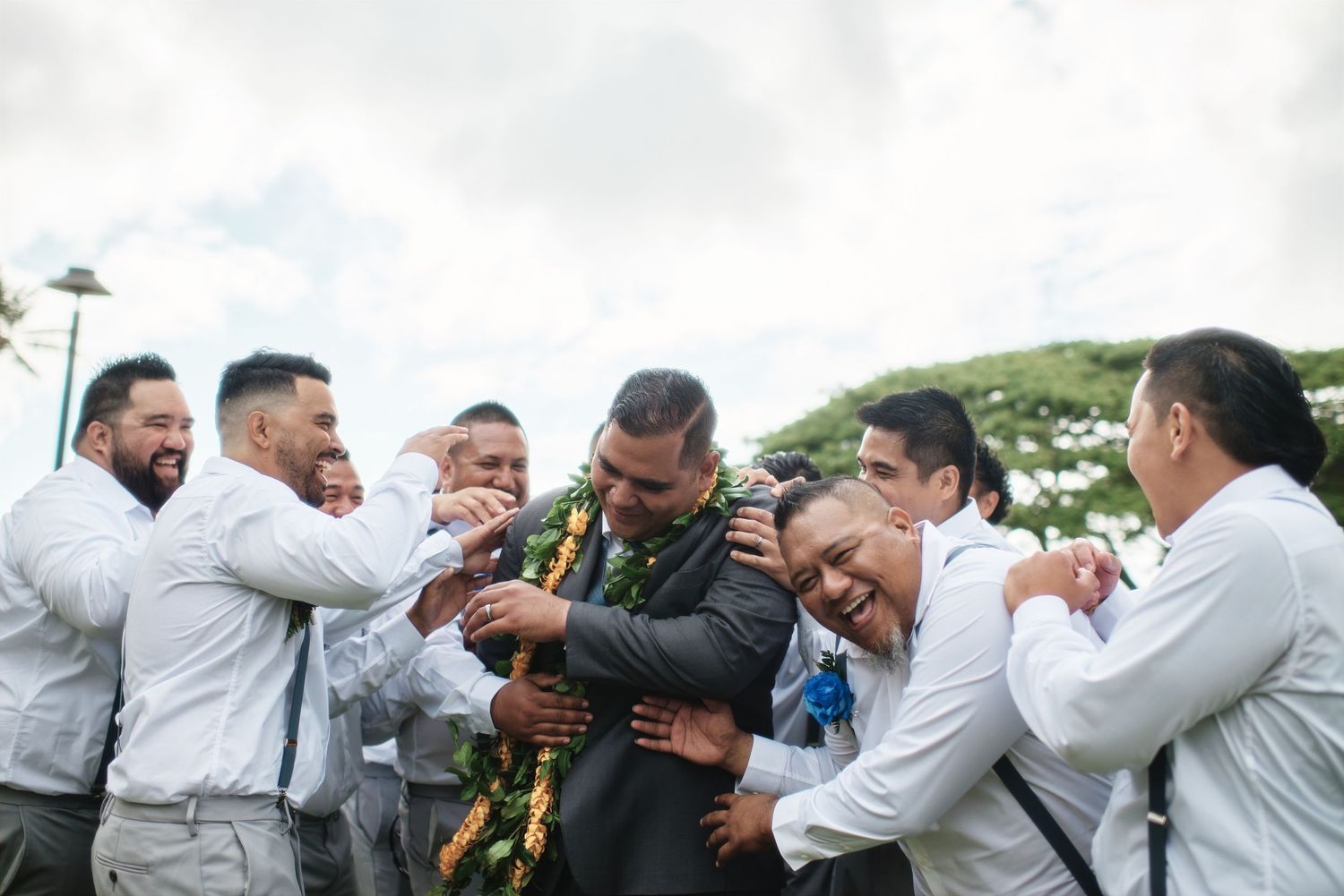Groomsmen in light gray suits celebrate and pose playfully for wedding photos outdoors.