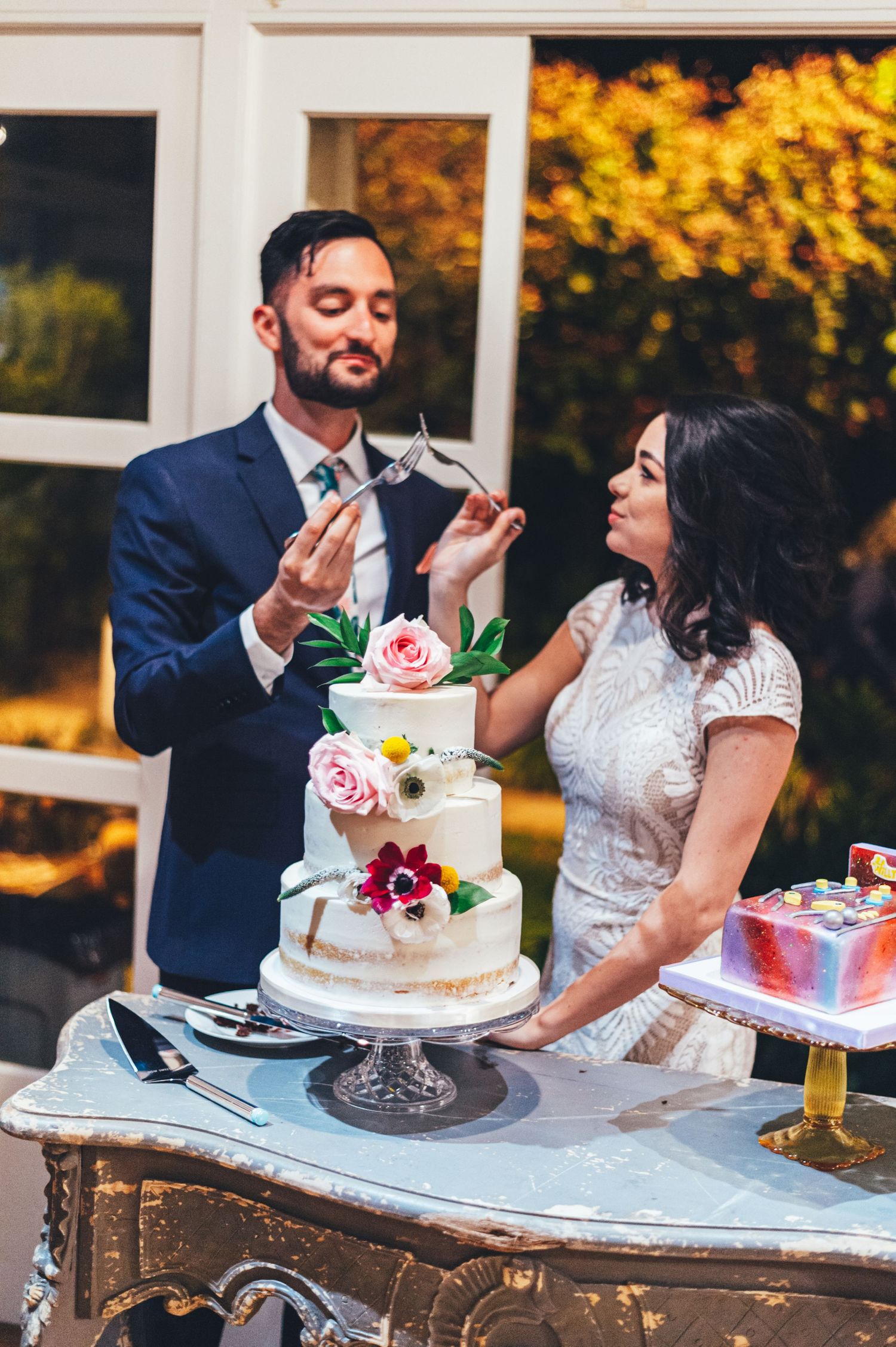 Wedding reception scene with couple sharing cake against backdrop of fall colors.