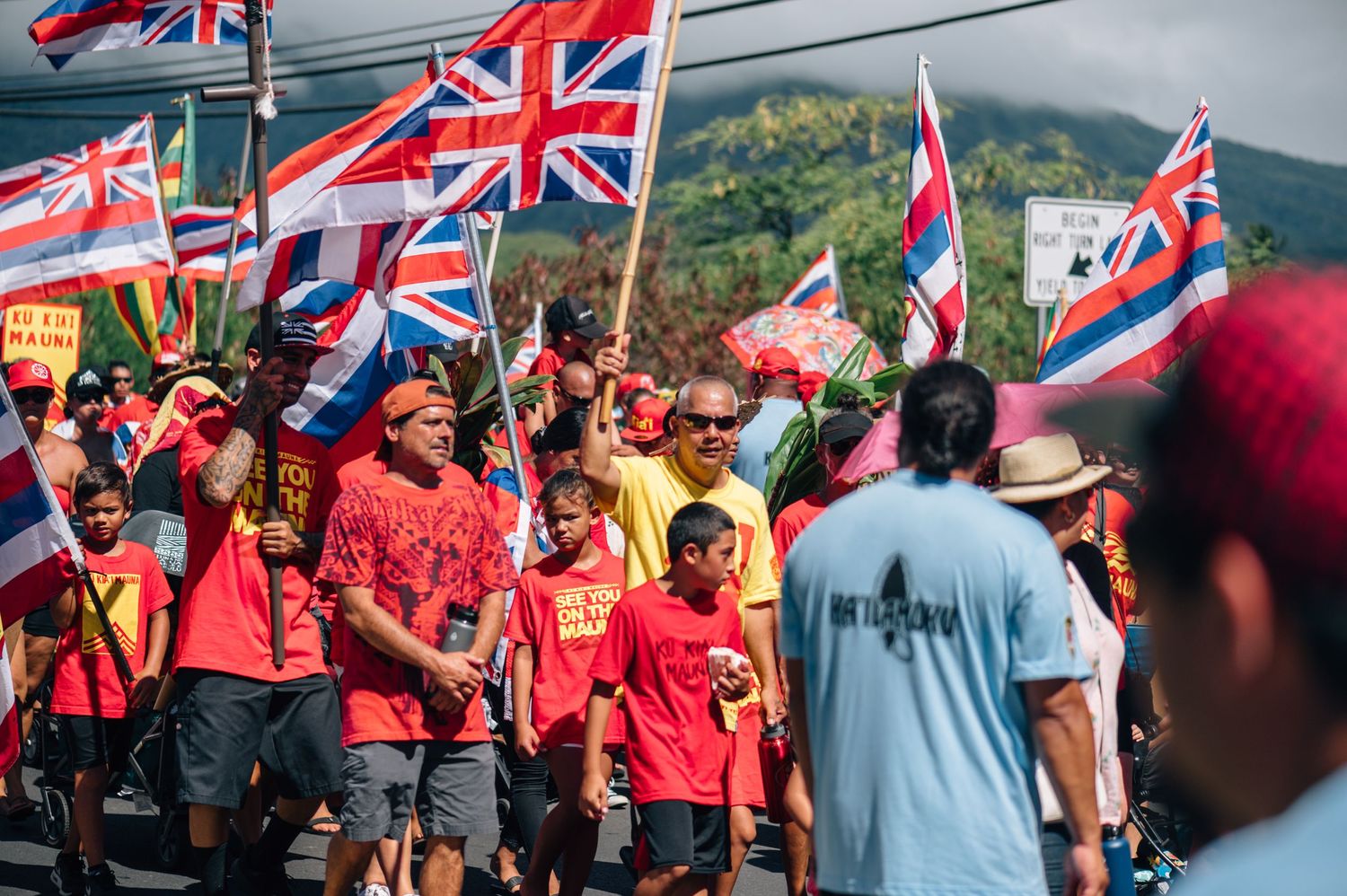 Protesters carrying Hawaiian and British flags gather for demonstration near mountain landscape.