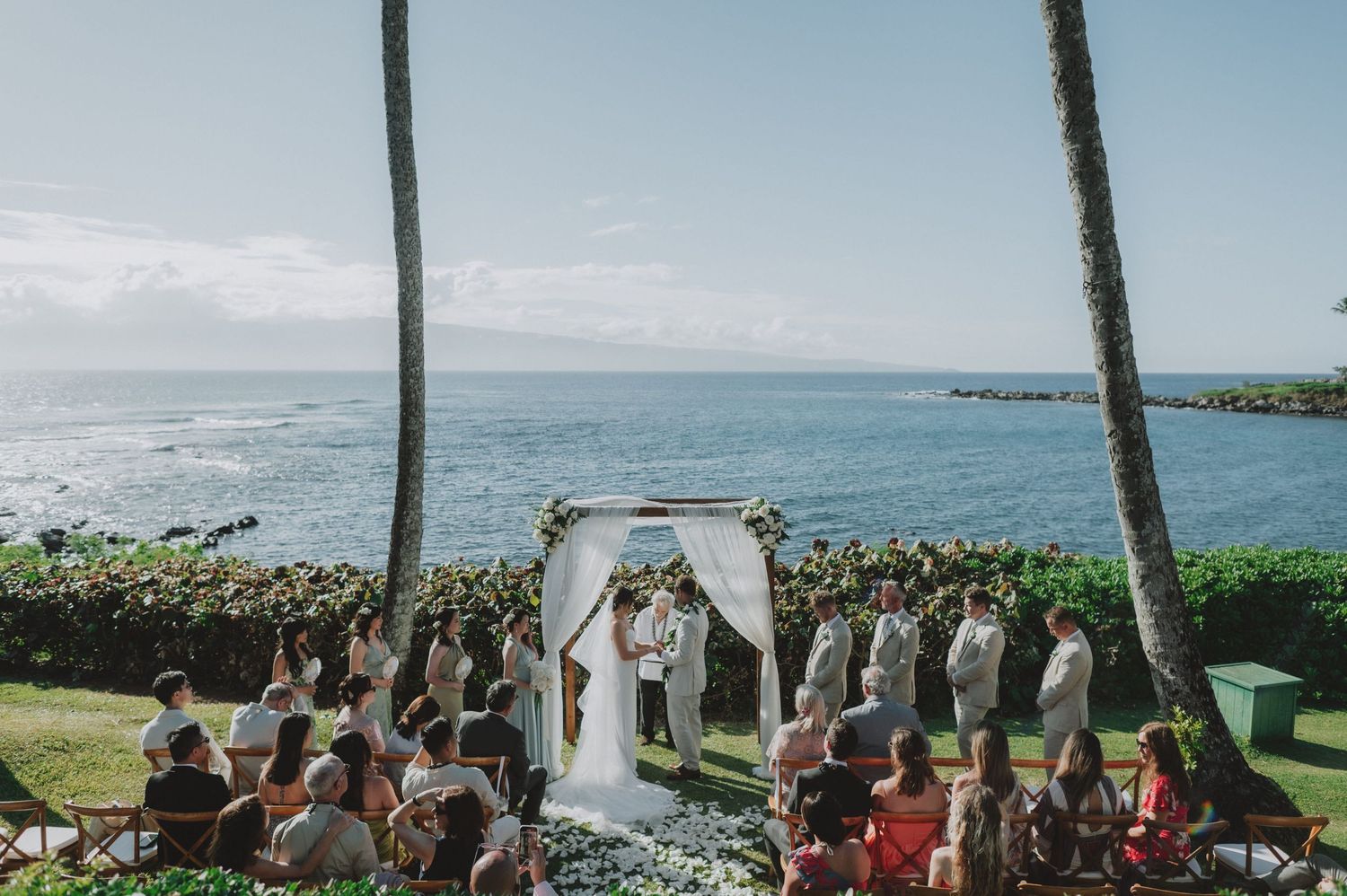 Outdoor Hawaiian wedding ceremony with ocean backdrop, guests seated, couple at floral arch between palm trees.