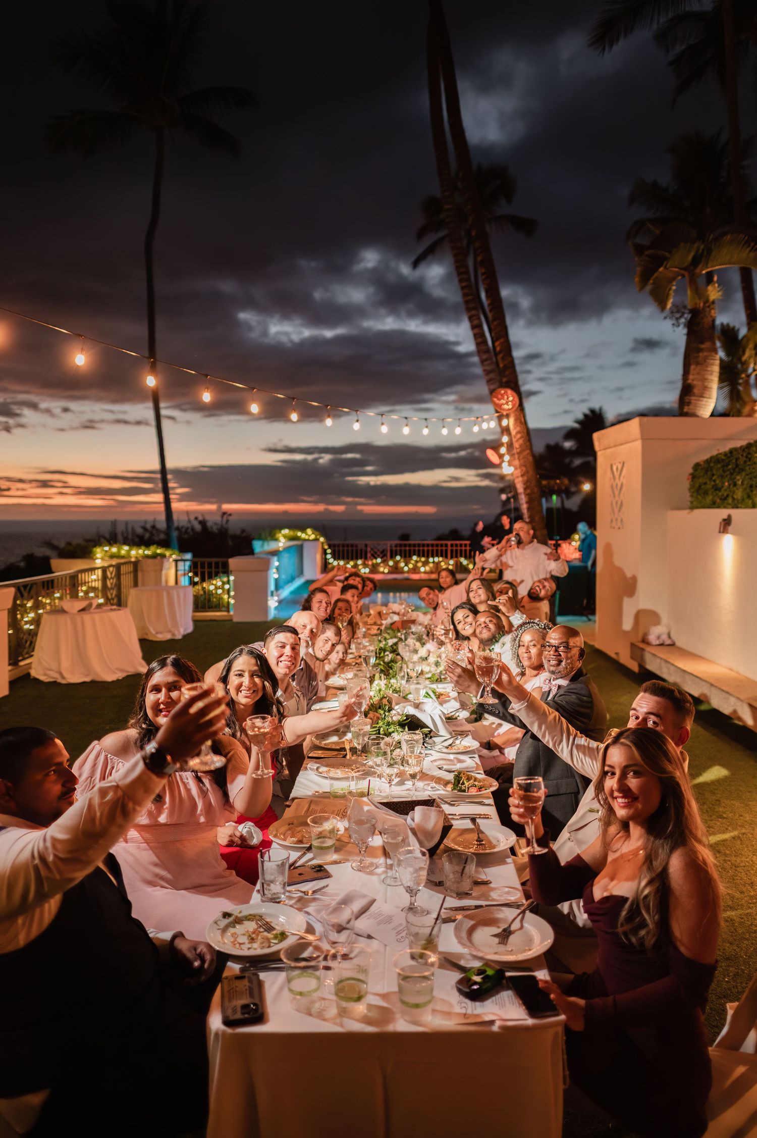 Guests toast at an elegant outdoor wedding reception dinner under string lights at sunset near the ocean.
