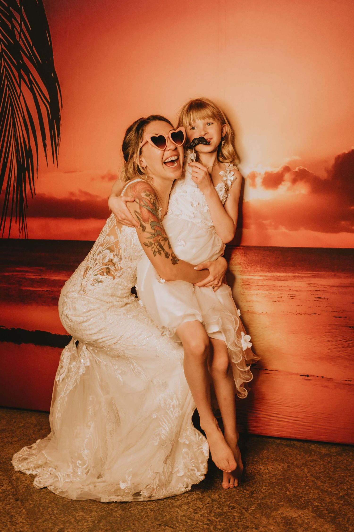 Bride in lace gown laughing with flower girl wearing heart sunglasses in front of tropical sunset backdrop.