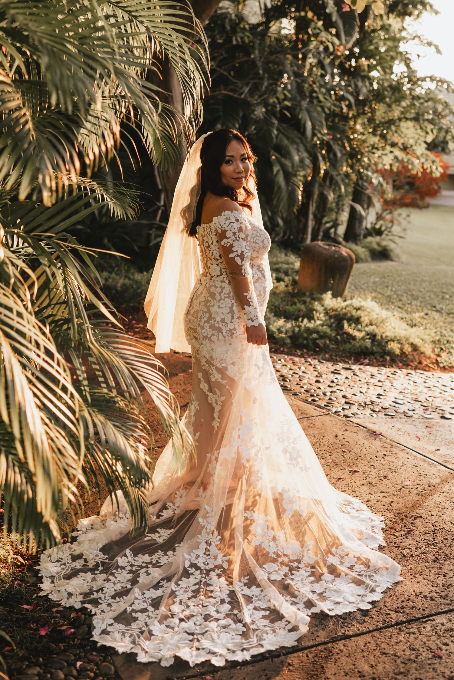 Bride in off-shoulder lace mermaid wedding gown with veil poses in tropical garden at golden hour.