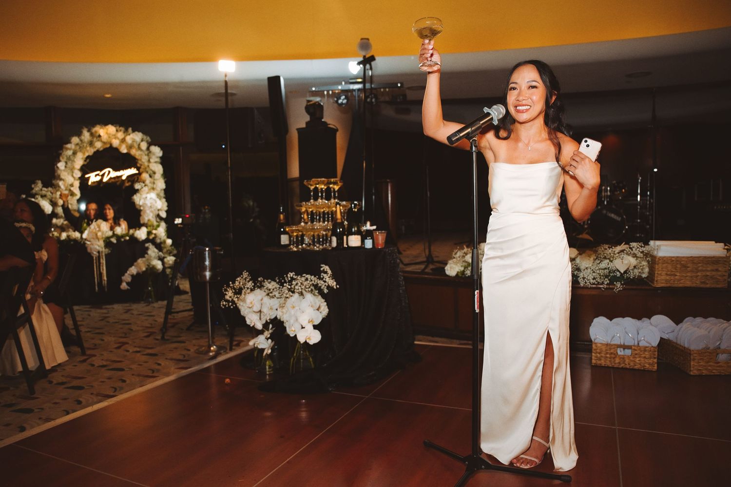 Bride in white gown raises champagne glass while giving speech at elegant wedding reception venue.