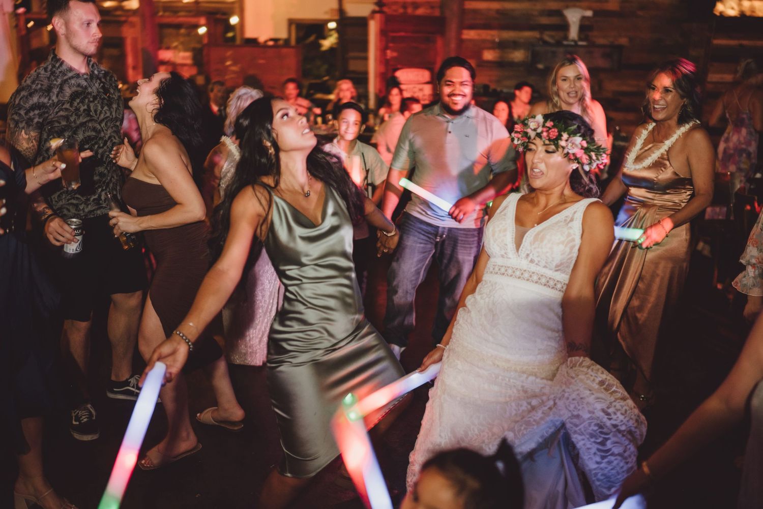 Two women dance joyfully at a wedding reception holding colorful glow sticks on a crowded dance floor.