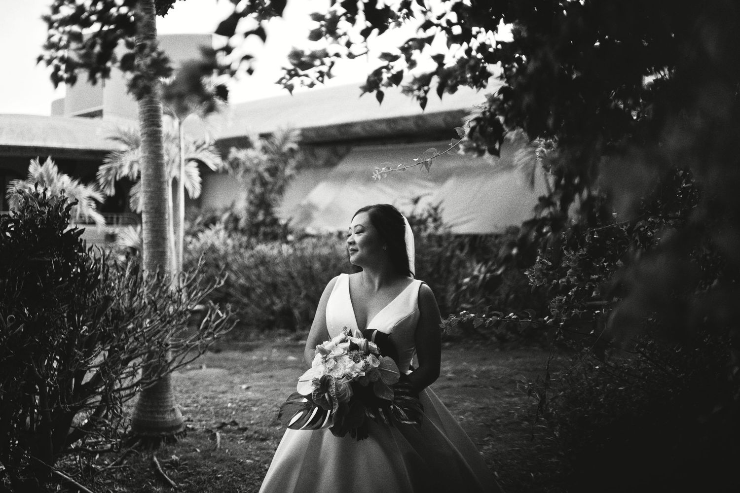 Black and white photo of a bride in a ball gown holding a bouquet, standing outdoors among lush trees and greenery.