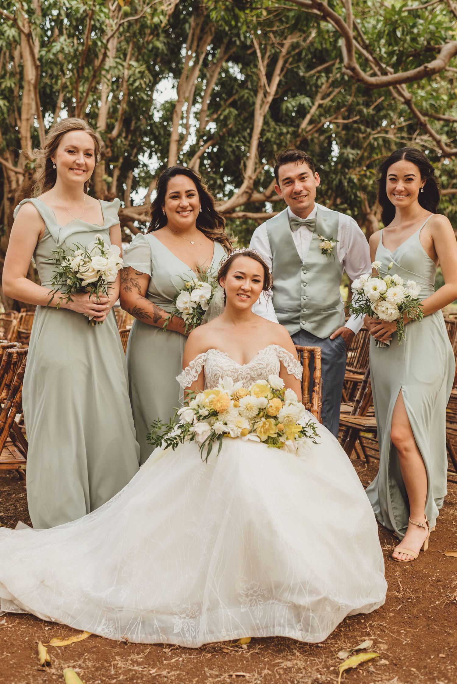 Bride in white gown sits surrounded by bridesmaids in sage green dresses and a groomsman at an outdoor wedding.