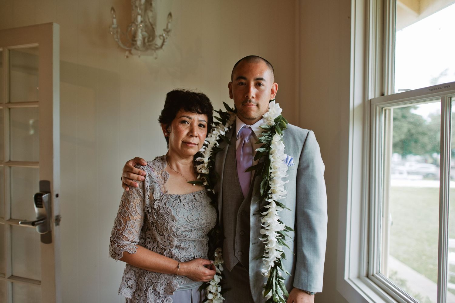 A groom in a gray suit with a floral lei poses with a woman in a lace dress at a wedding venue.