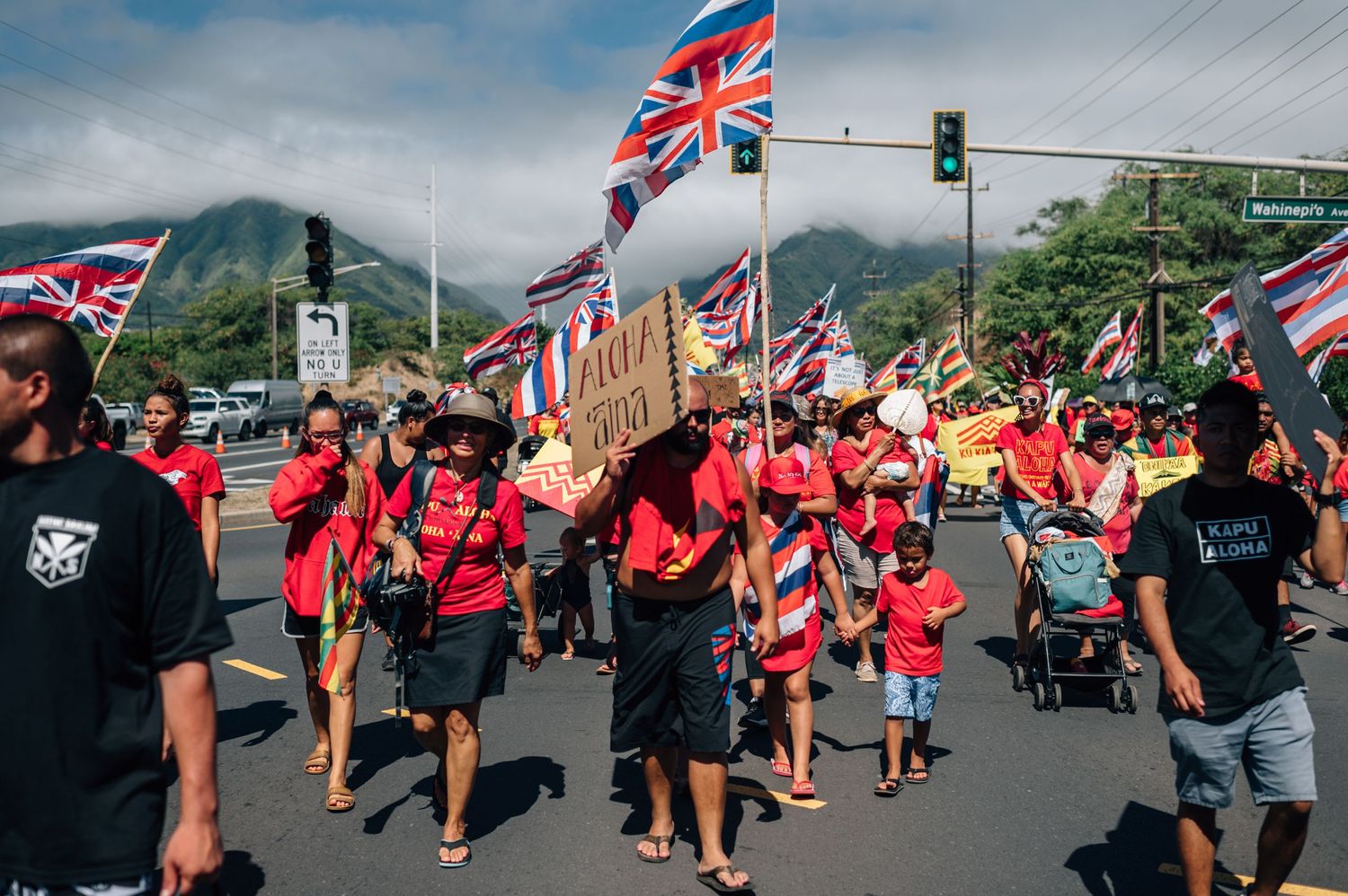 Large crowd marches with Hawaiian state flags and protest signs on a sunny day with mountains visible.