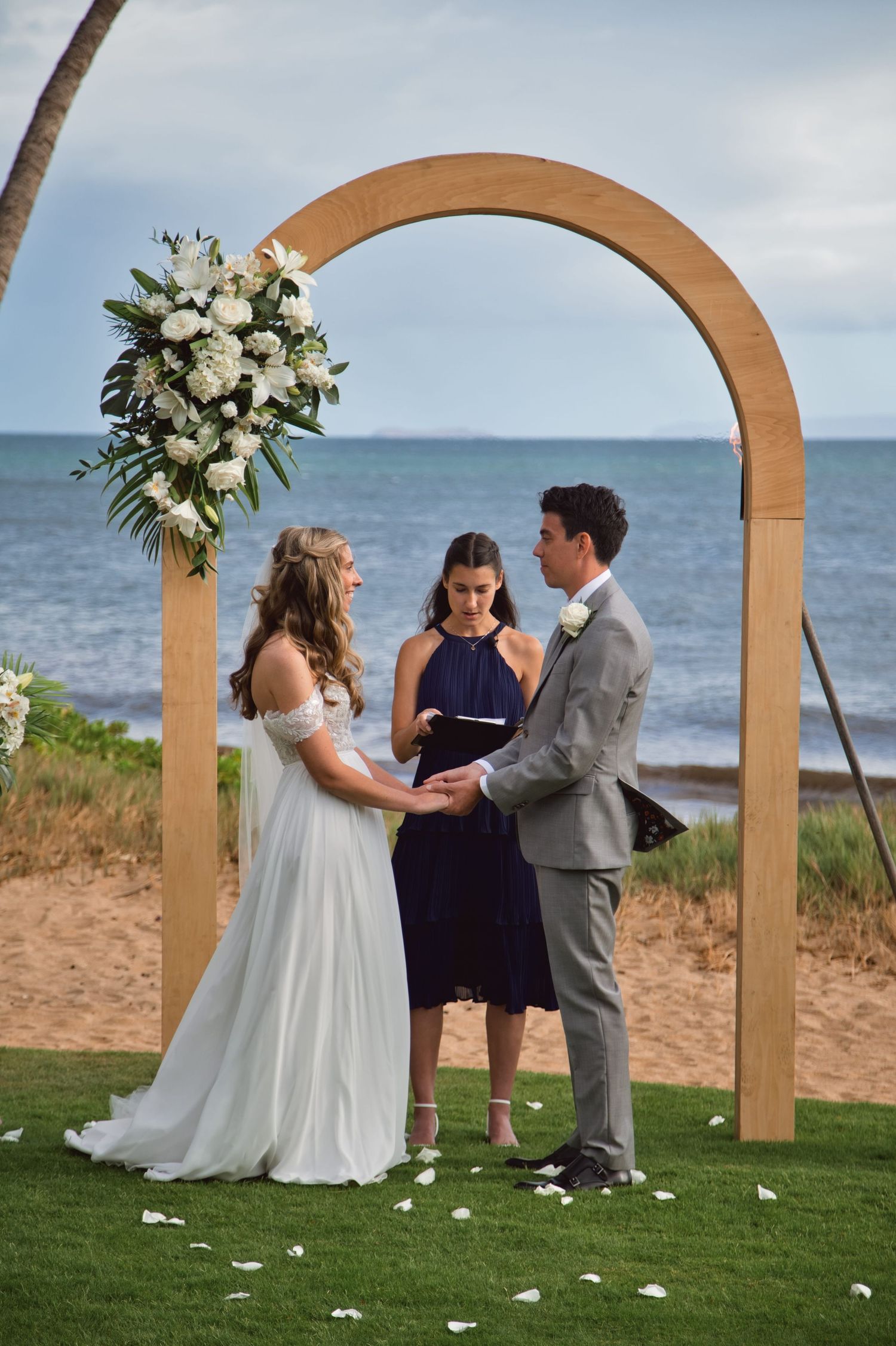 Couple exchanging vows under wooden arch with white floral arrangement on tropical beach with ocean backdrop.