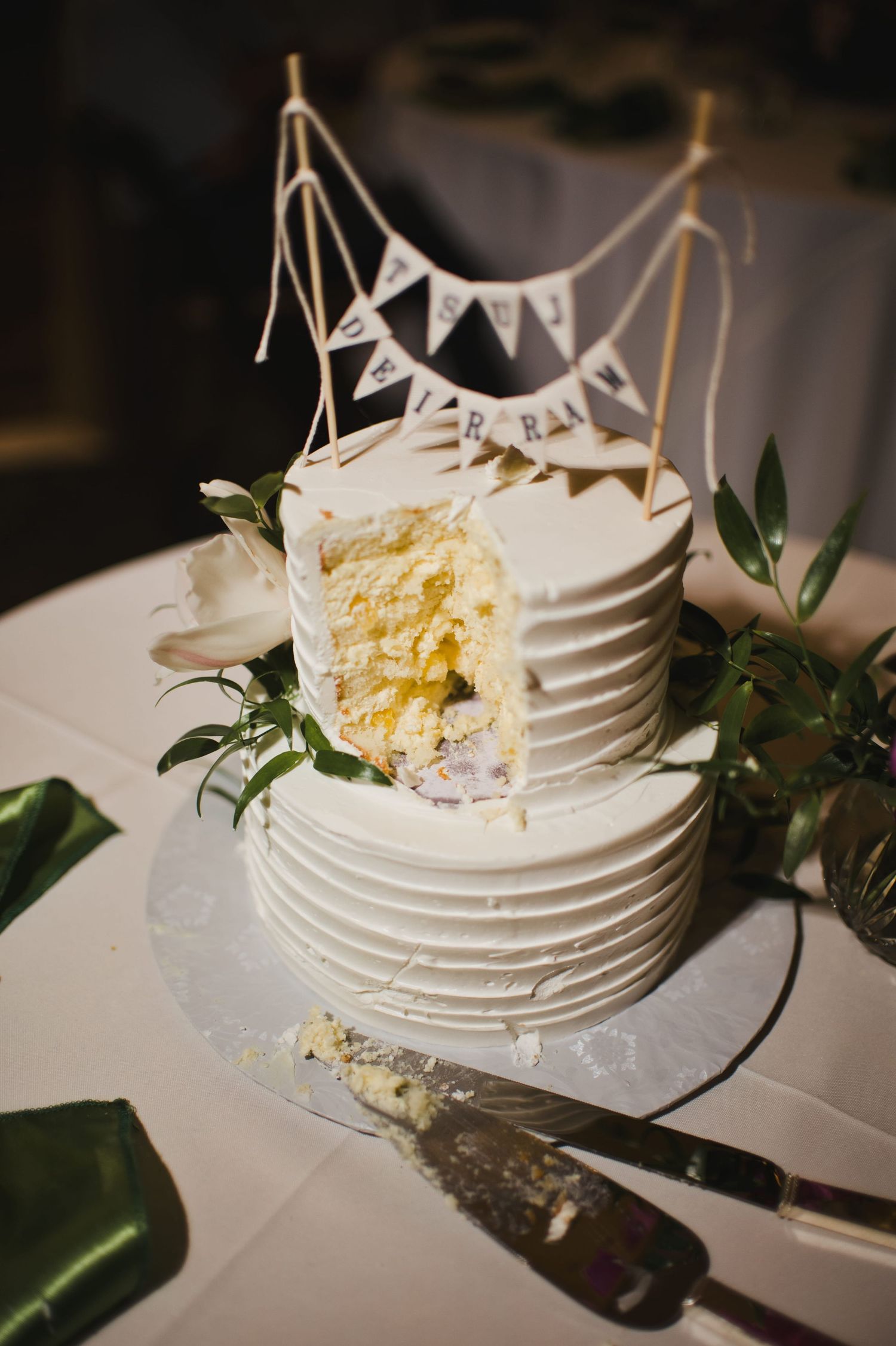 Two-tier white wedding cake with rustic bunting topper, greenery, and yellow interior slice revealed on white table.
