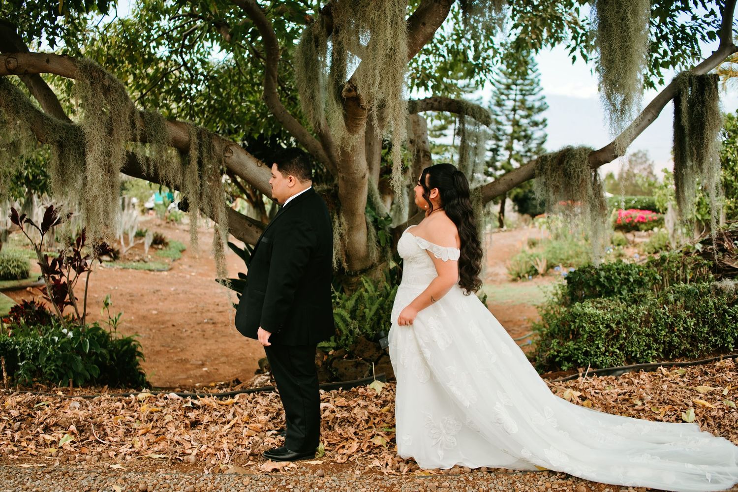Bride in white off-shoulder gown and groom in black suit share first look beneath a large tree in a nearby garden.