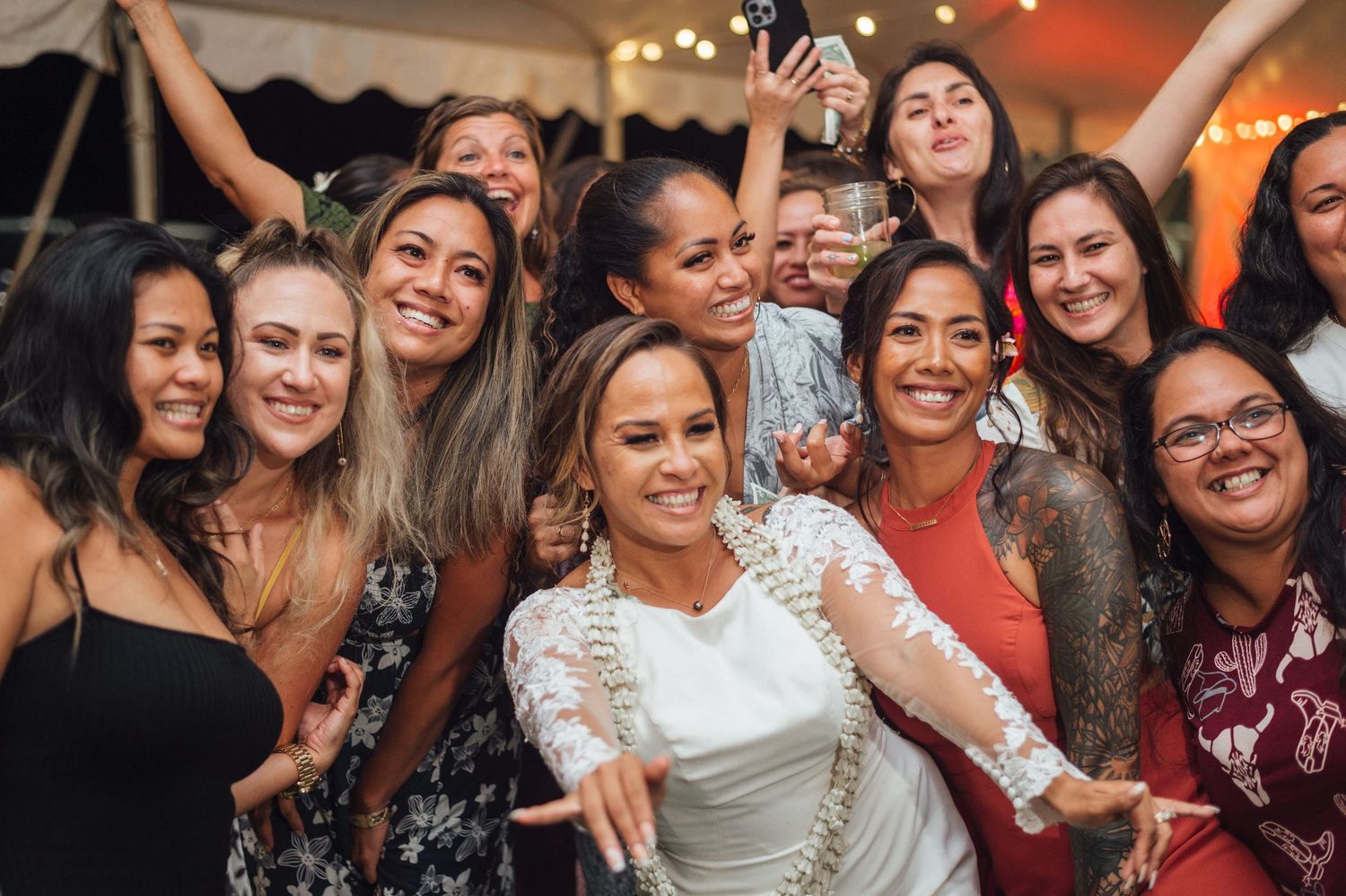 Bride in white lace dress smiling with group of women friends at wedding reception celebration.