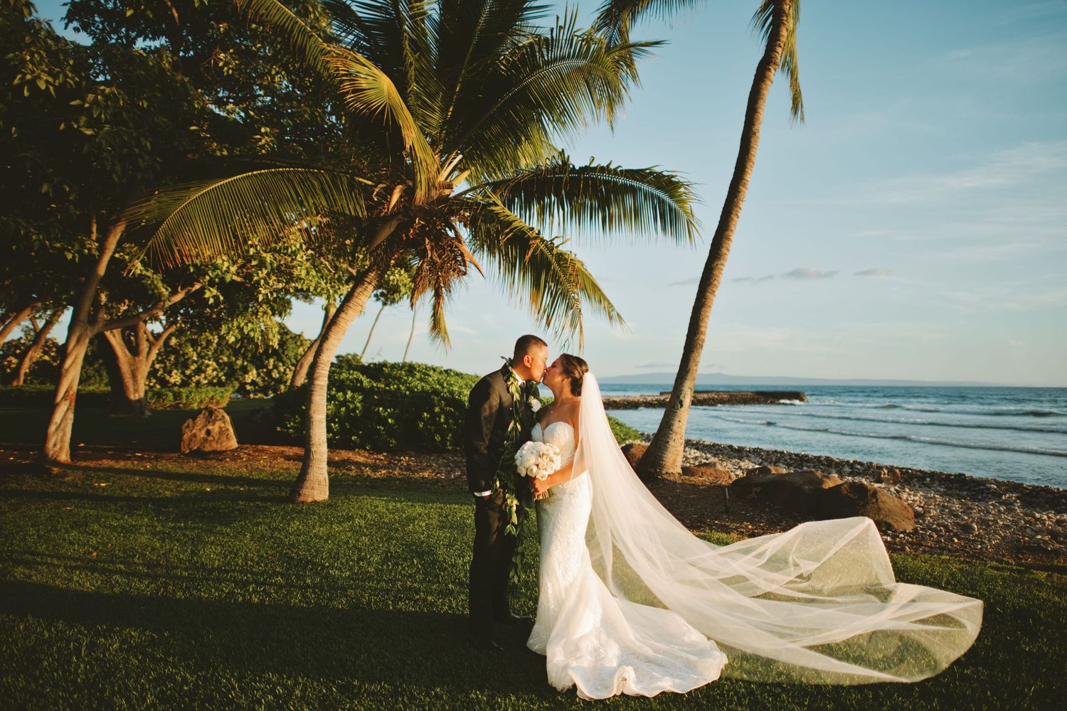 Bride and groom kissing under palm trees by the ocean, bride's veil flowing in the breeze at a tropical beach wedding.