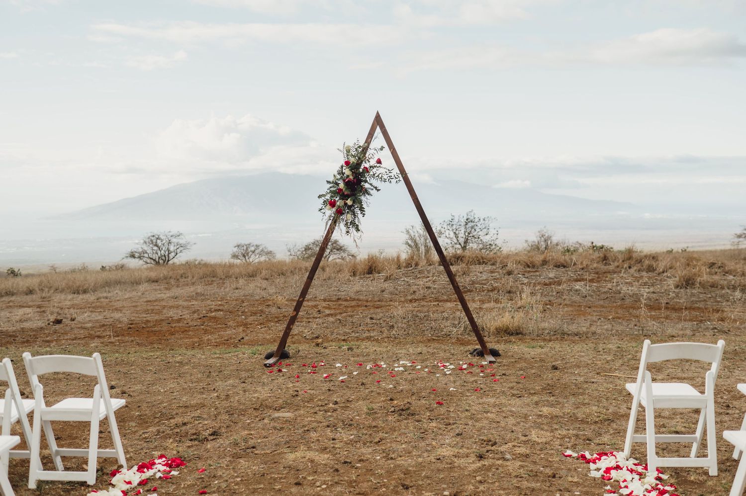 Rustic triangular wooden wedding arch with floral accents set outdoors on a Hawaii hilltop with mountain views.