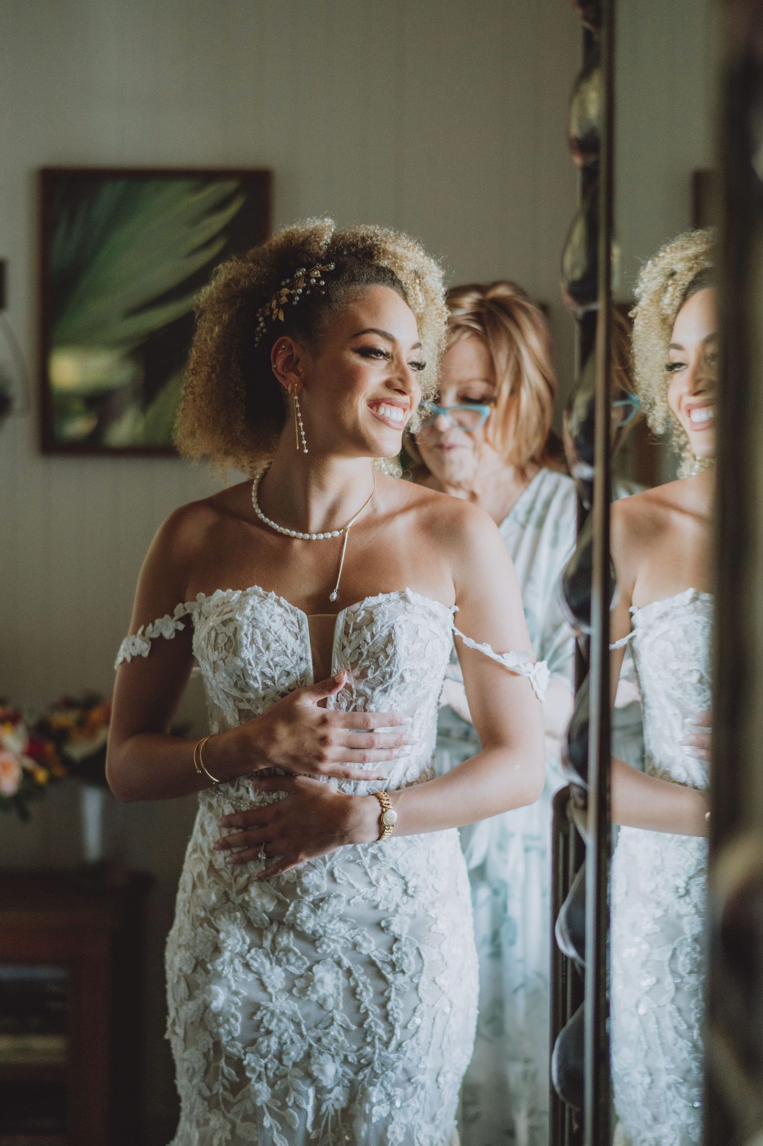 Bride in lace off-shoulder wedding gown smiling at mirror reflection, wearing pearl jewelry and floral headpiece.