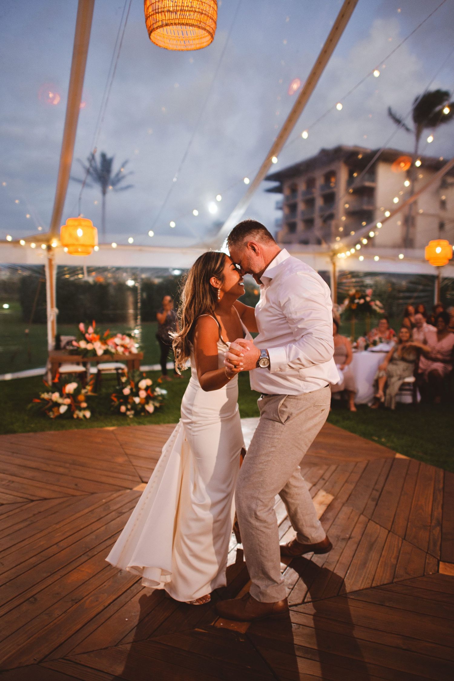 Couple sharing first dance under string lights on wooden dance floor at elegant outdoor wedding reception at dusk.
