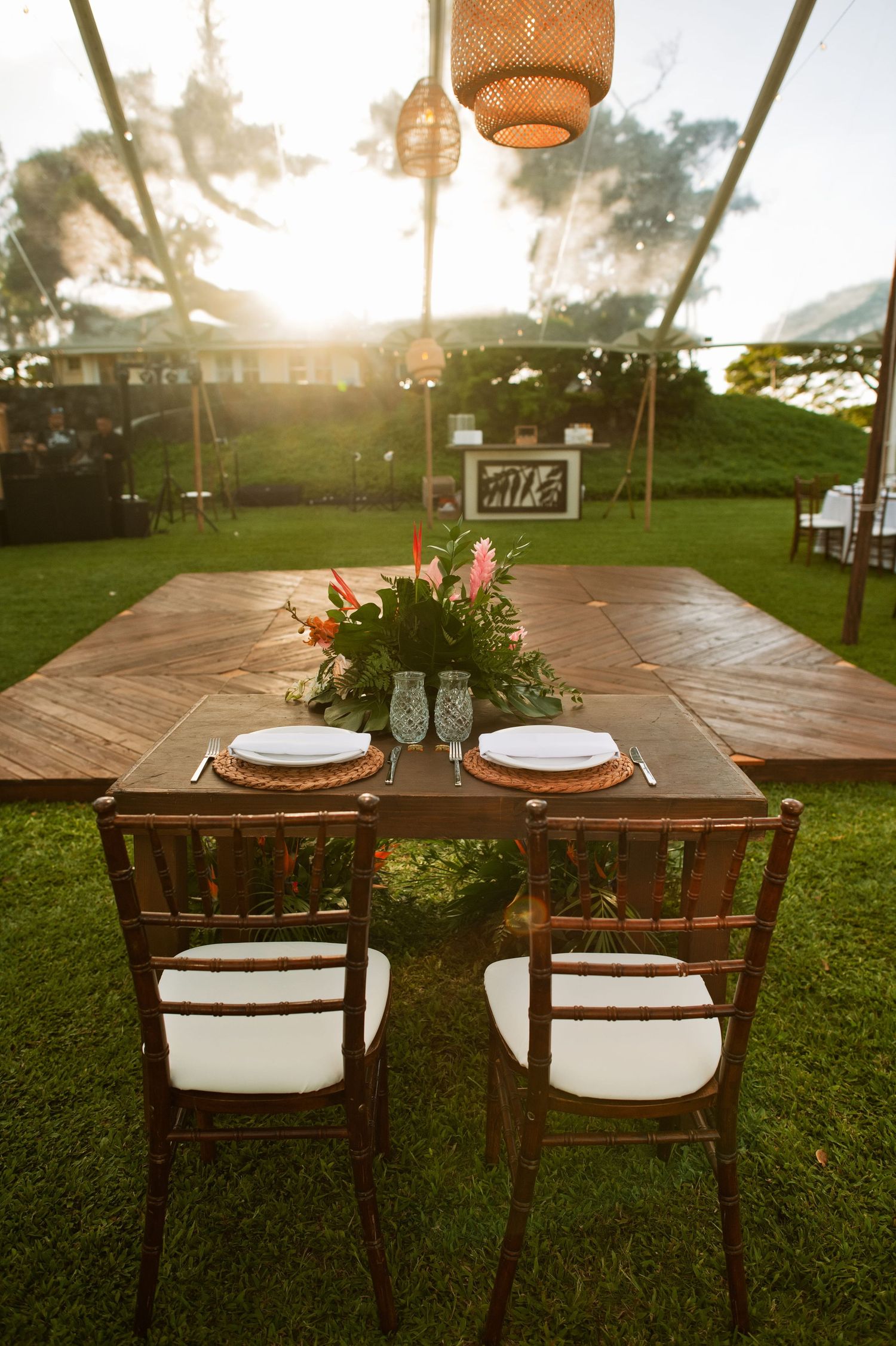 Elegant outdoor wedding sweetheart table with wooden chairs, tropical floral centerpiece, and warm sunset lighting under a tent.