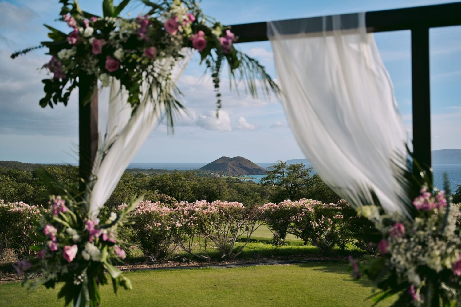 Elegant outdoor wedding arch adorned with pink flowers and white draping, overlooking lush gardens and coastal hills.