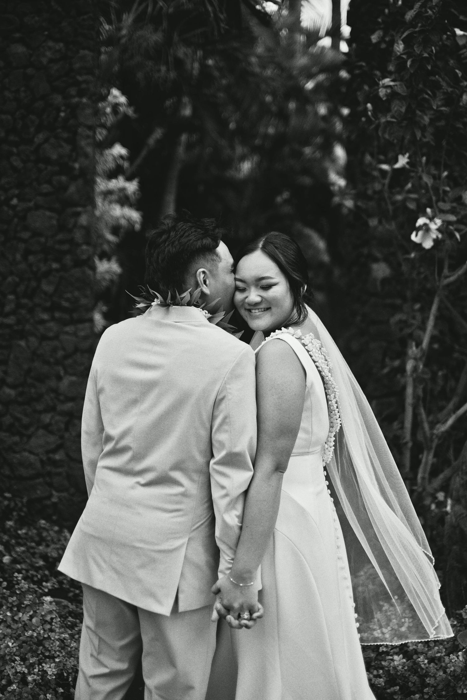 Black and white photo of a smiling bride in veil holding hands with groom in suit sharing a kiss outdoors.