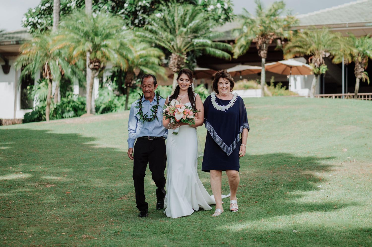 Wedding ceremony procession on grassy lawn with white arbor and palm trees.