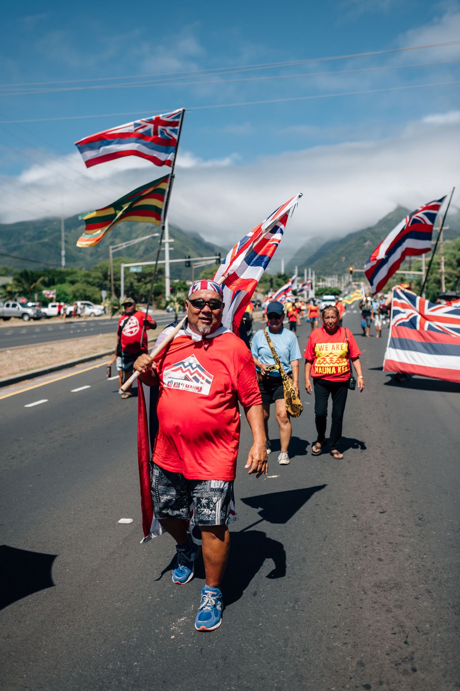 Demonstrators wave flags and march along a road with volcanic mountains visible in the distance.