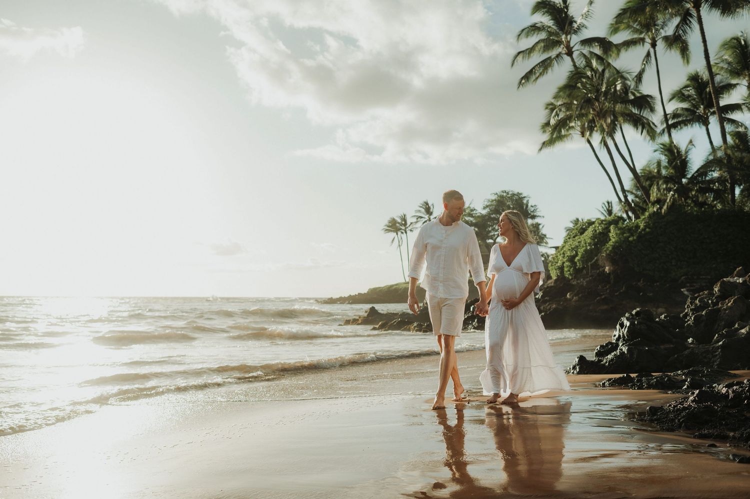 Pregnant couple in white walking hand-in-hand on a tropical beach at golden hour with palm trees and ocean waves.