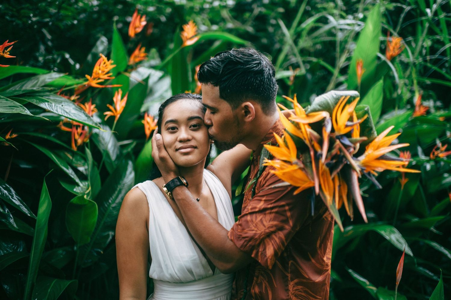 Couple in tropical garden with bird of paradise flowers, man kissing woman's cheek, romantic outdoor portrait.