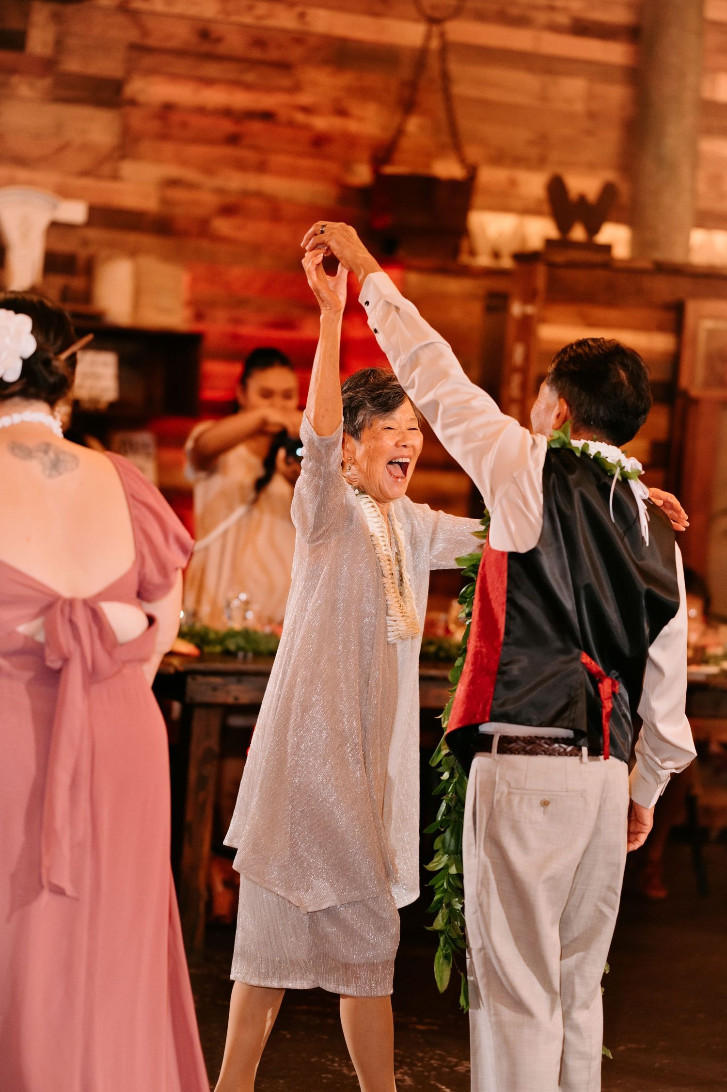 Elderly couple dancing joyfully at a rustic wedding reception, raising hands together in celebration.