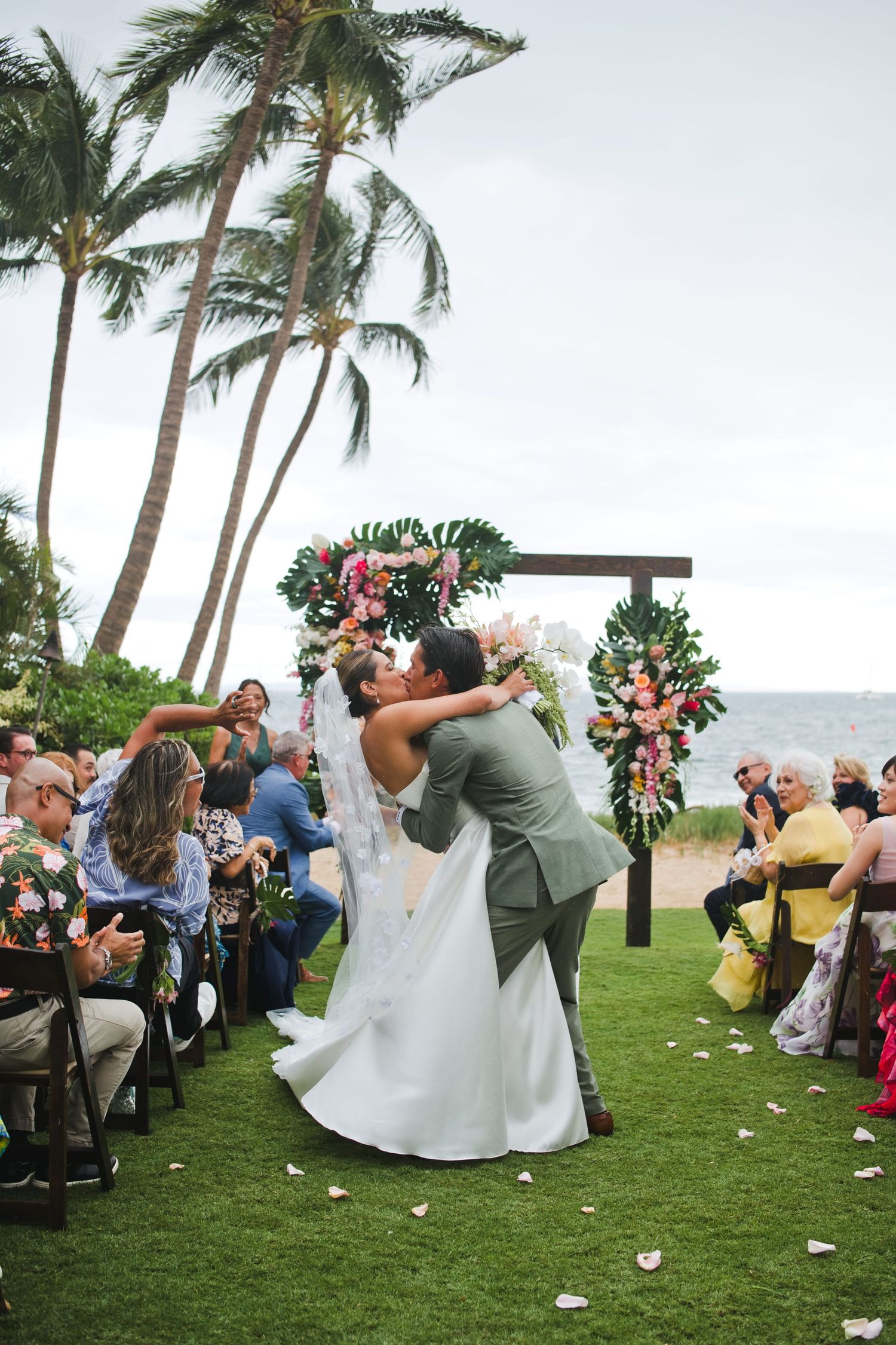 Couple sharing first kiss at tropical outdoor wedding ceremony with floral arch, palm trees, and ocean backdrop in Maui.