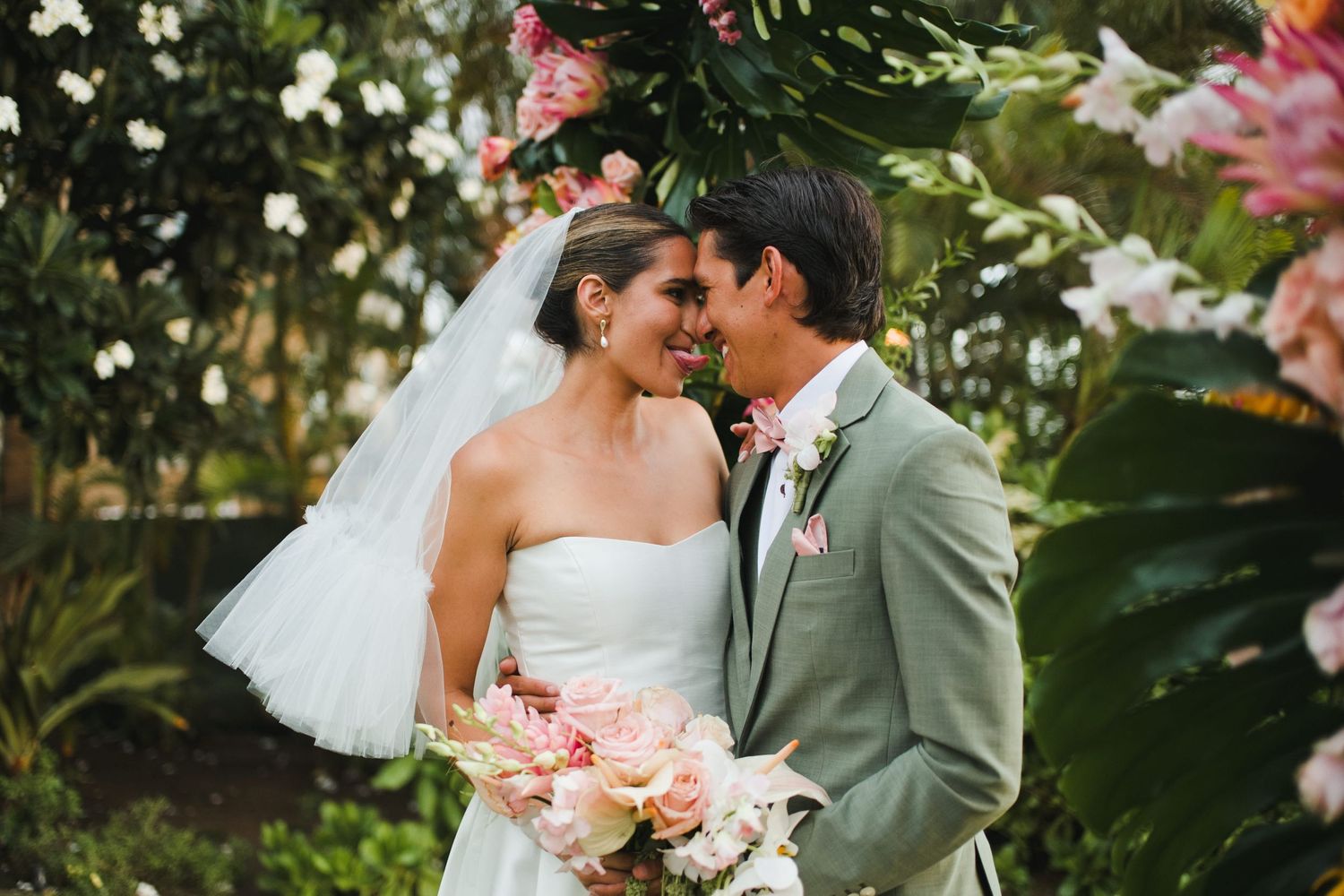Bride and groom share a romantic moment outdoors, surrounded by lush tropical flowers and greenery on their wedding day.