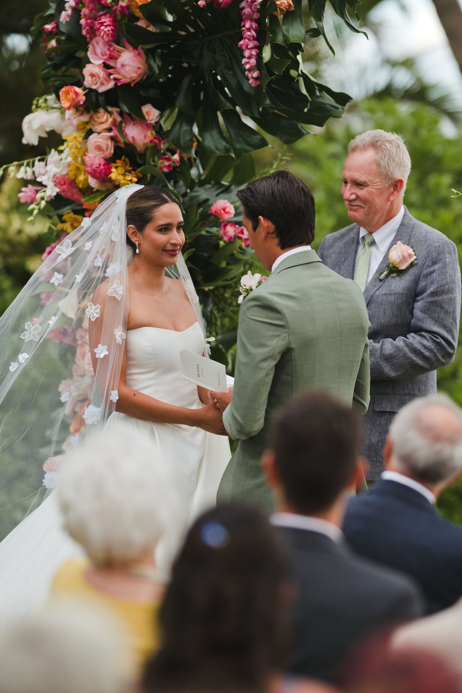 Bride and groom exchange vows at outdoor wedding ceremony beneath colorful floral arch with officiant.