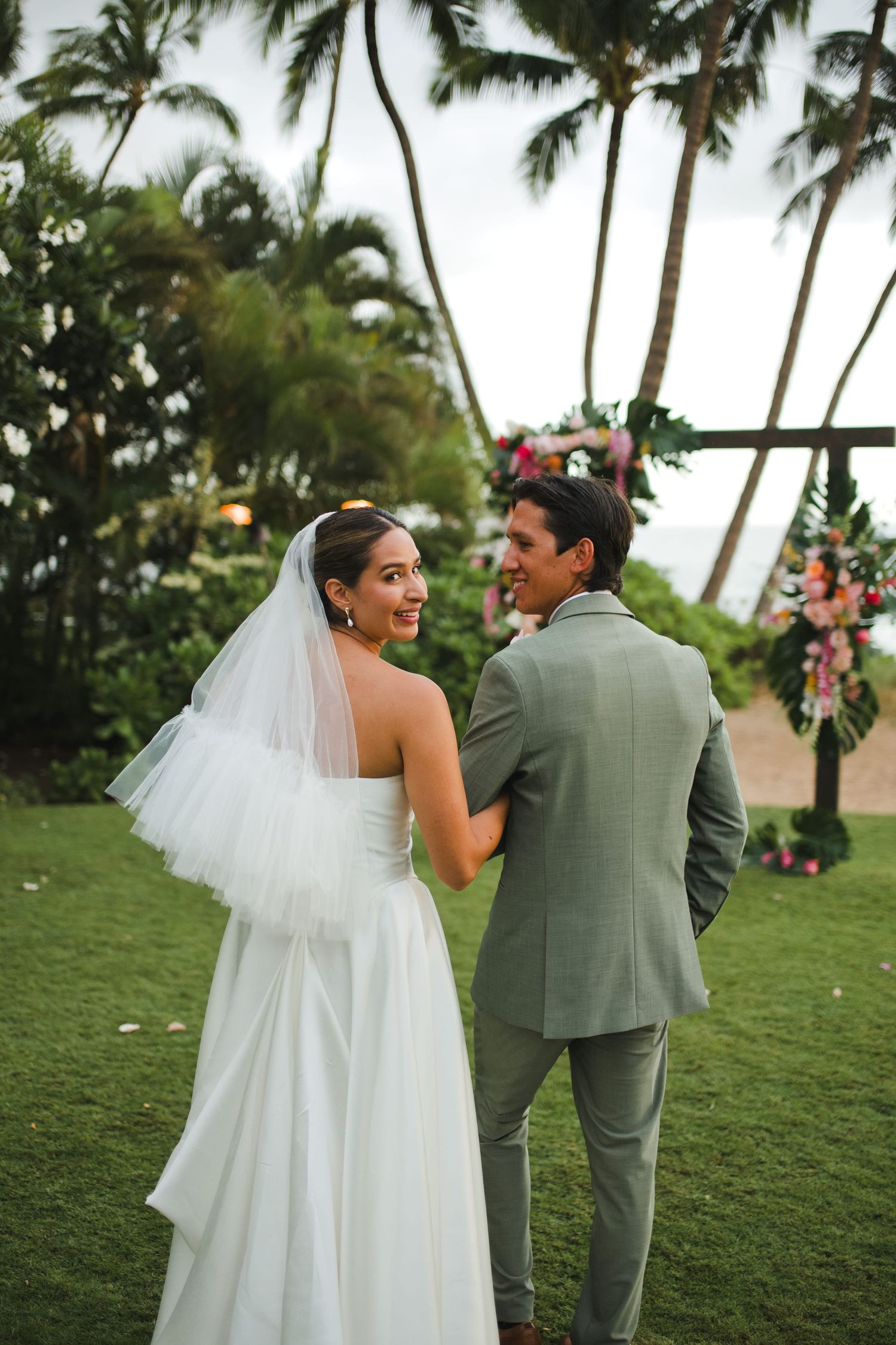 Bride in white gown and veil stands with groom in gray suit at tropical outdoor wedding ceremony with palm trees.