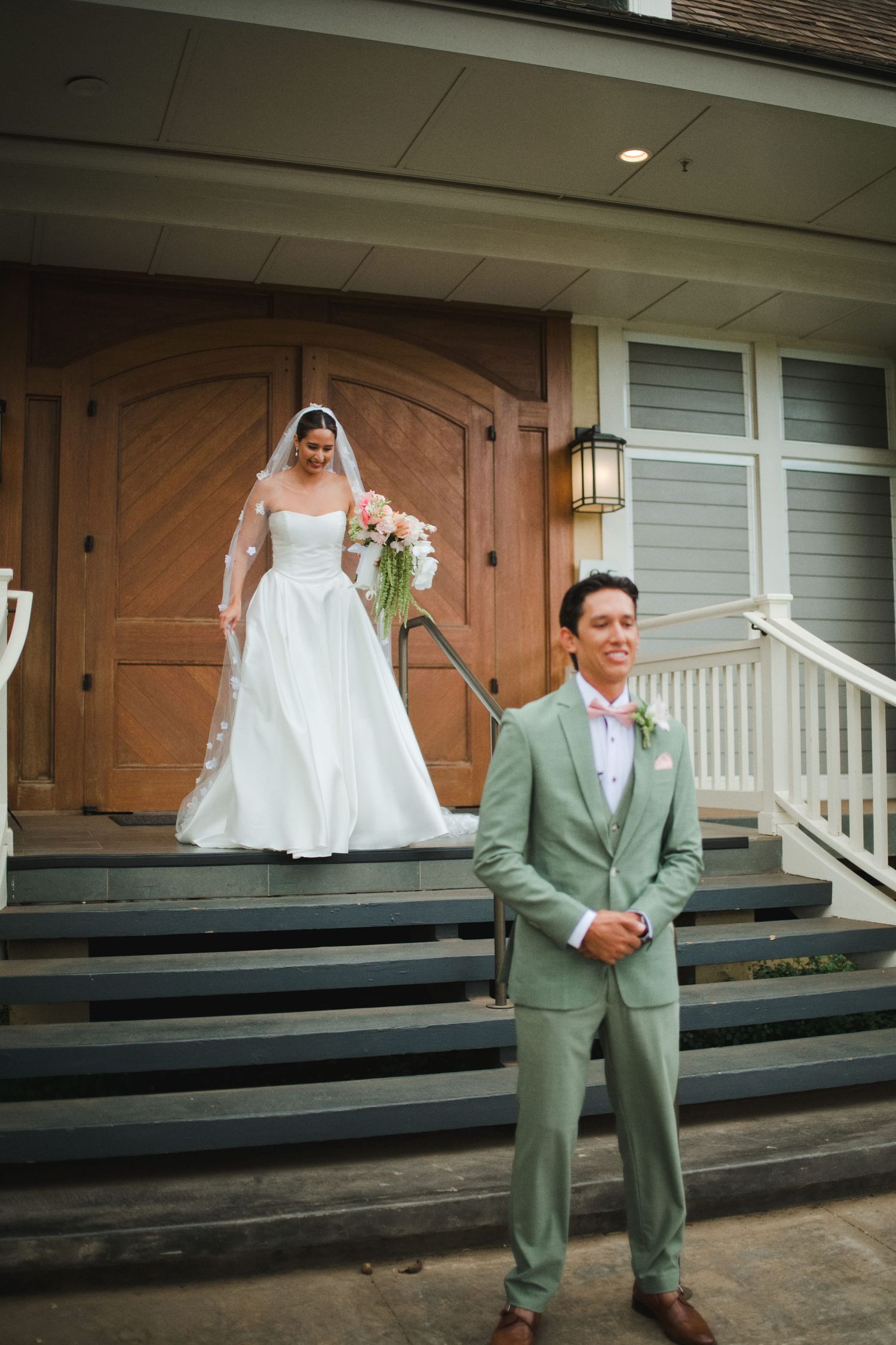 Groom in sage green suit stands on steps as bride in white gown with veil descends behind him at wedding venue.