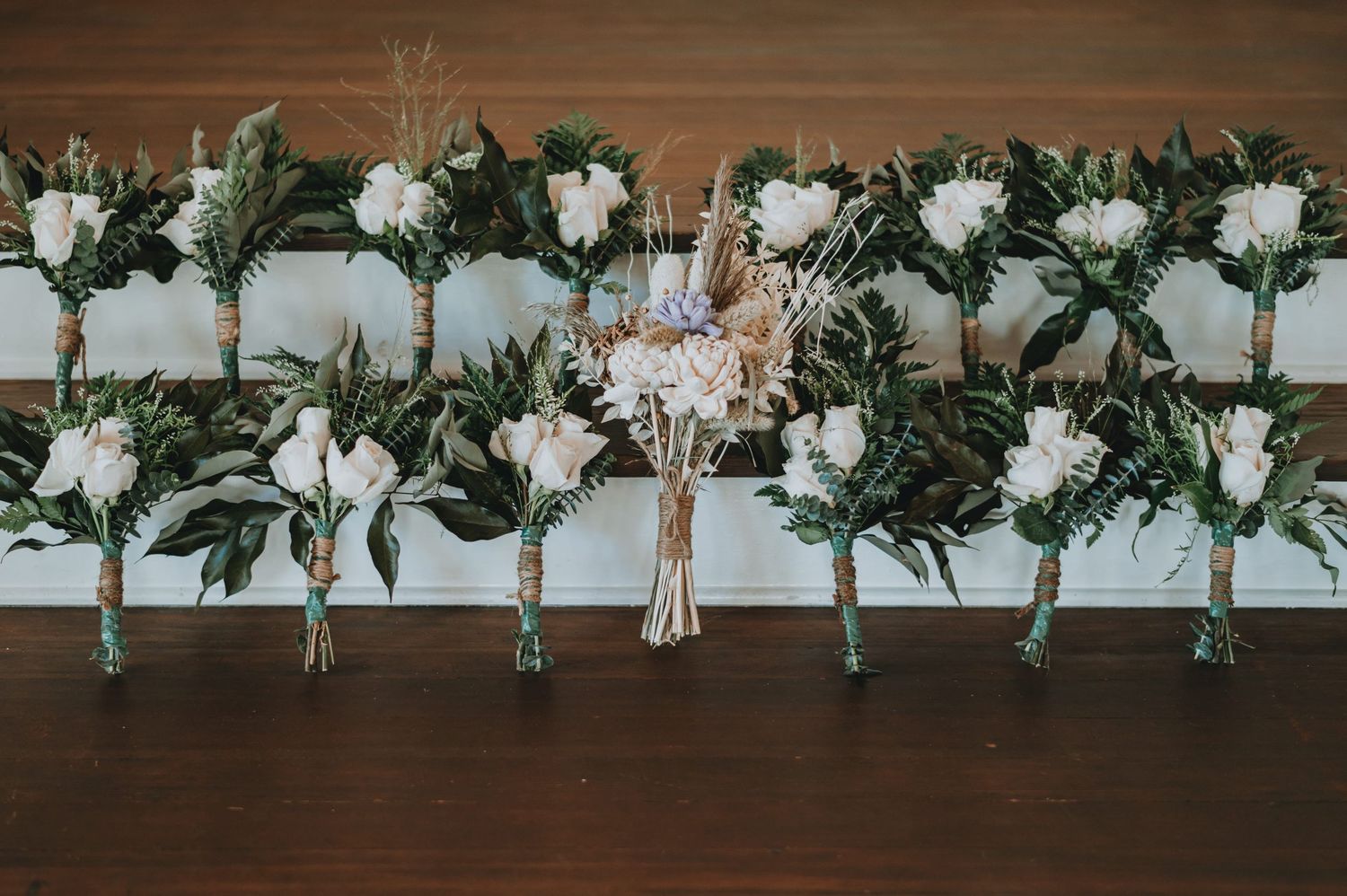 Row of white floral boutonnieres with greenery and dried flowers displayed on a rustic wooden surface.