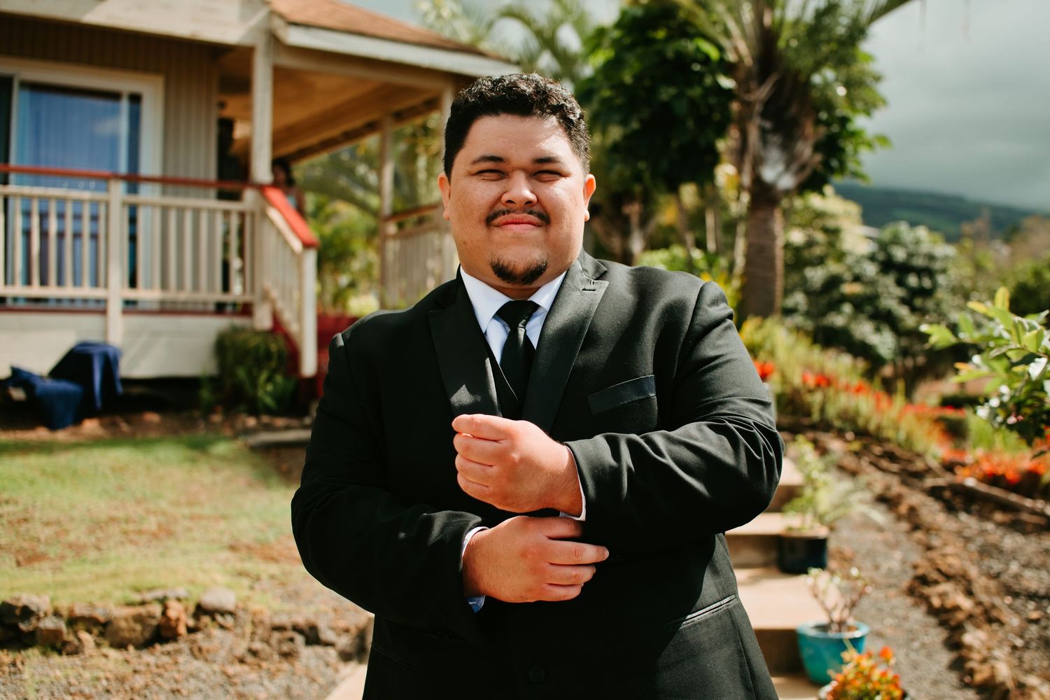 Young man in black suit adjusting cufflinks outdoors near a house with tropical garden background.
