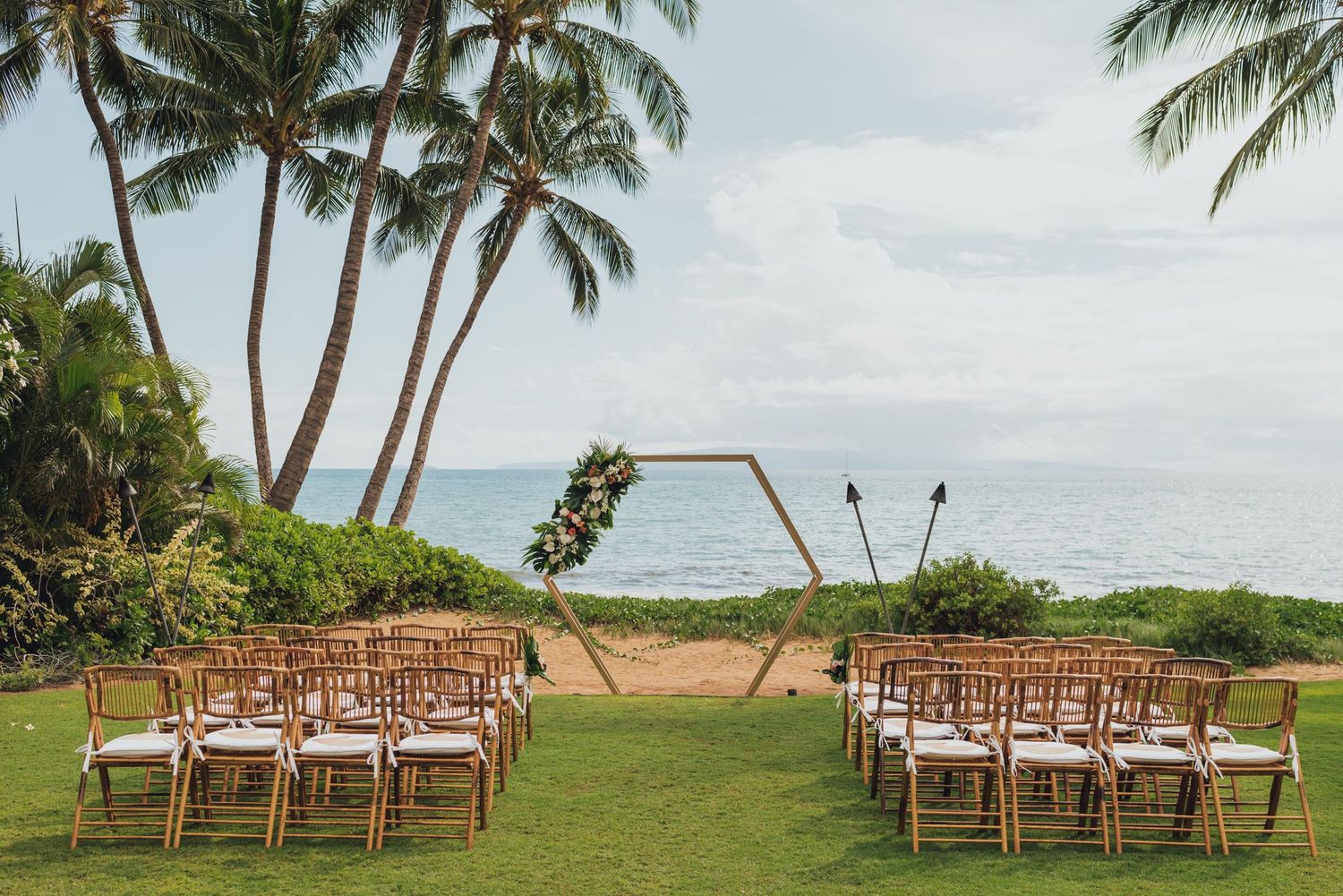 Outdoor beachside wedding ceremony setup with wooden chairs and floral arch framed by palm trees overlooking the ocean.