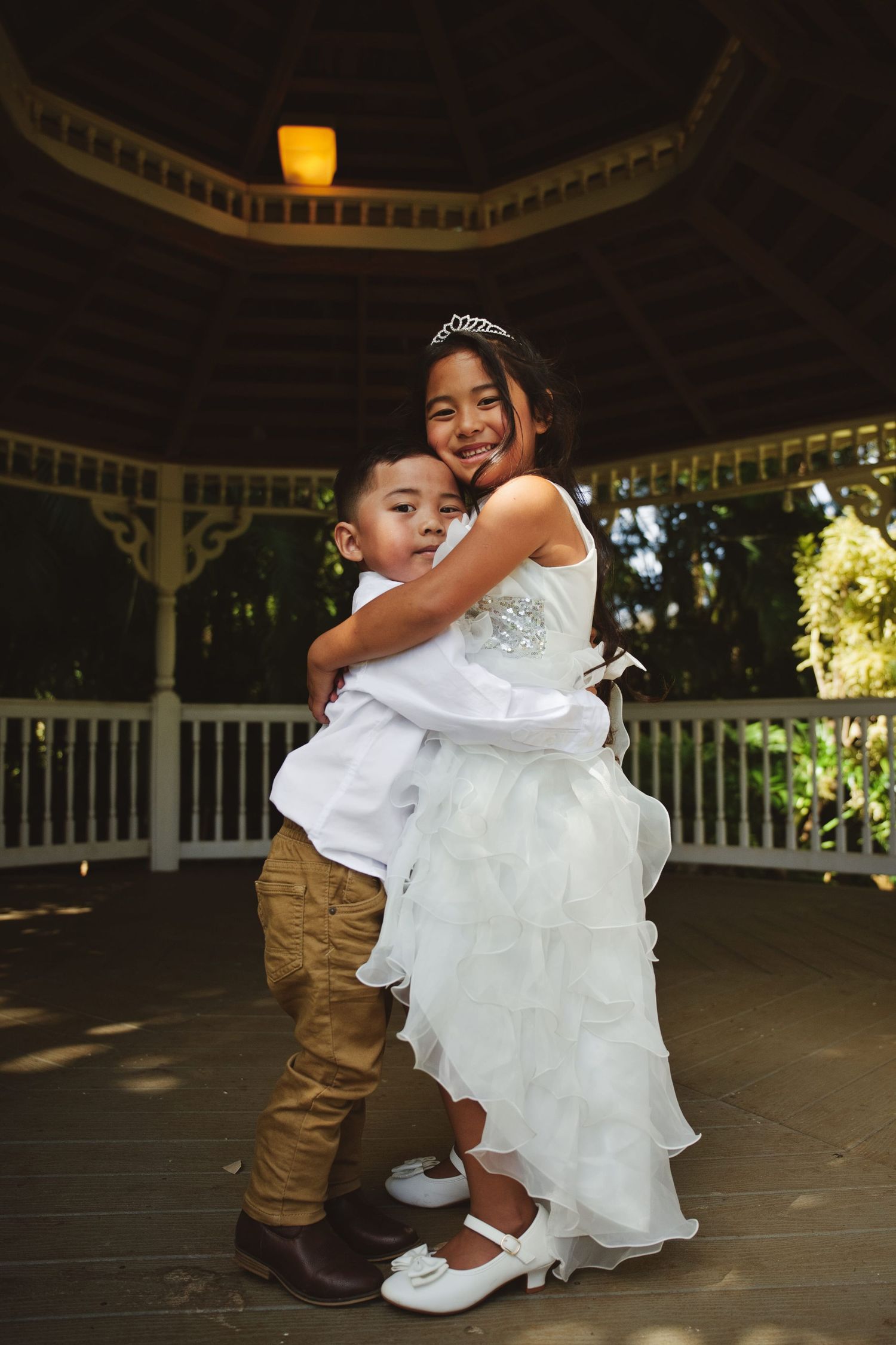 Two children in wedding attire hug in a gazebo, girl in white dress with tiara.