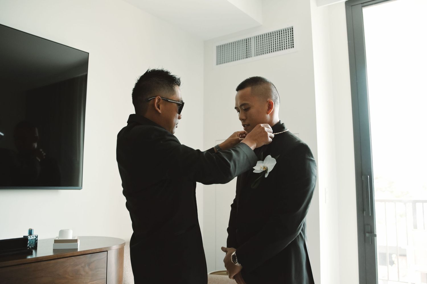 A man in sunglasses adjusts a boutonniere on a groom in a black suit before a wedding ceremony.