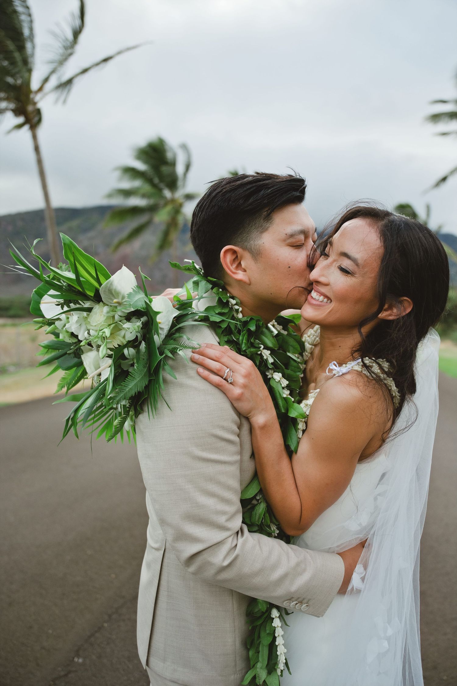 A groom in a beige suit kisses his smiling bride holding a tropical white bouquet with lush greenery in Hawaii.