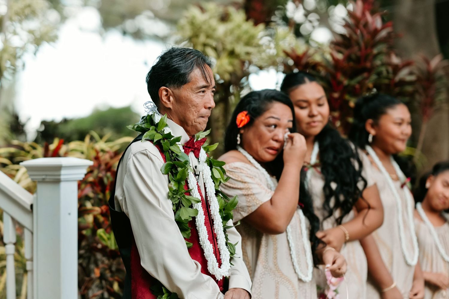Hawaiian wedding ceremony with guests in traditional leis and floral attire standing outdoors among tropical plants.