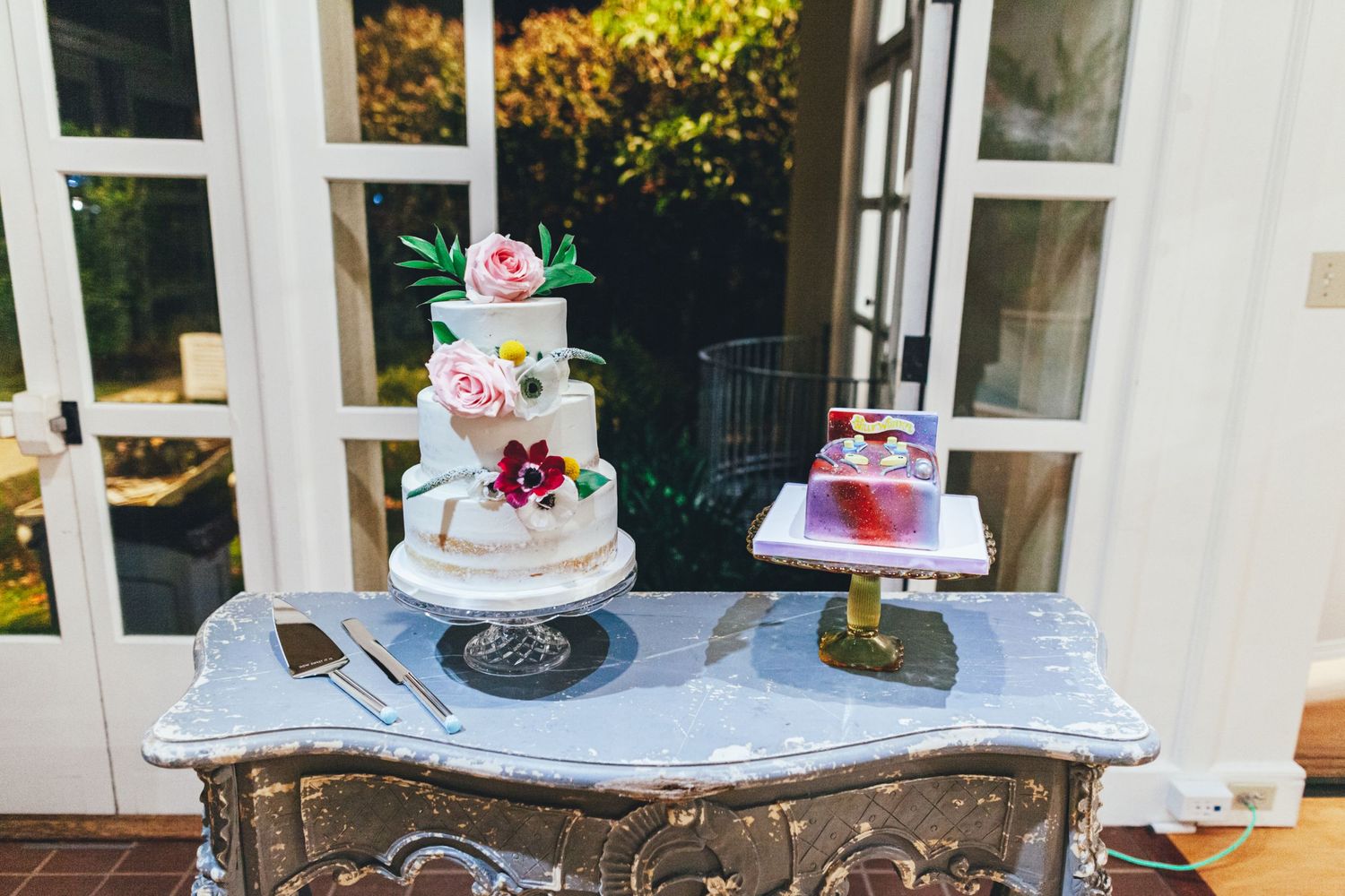 Three-tiered white wedding cake with floral decorations displayed on an antique marble-topped table.