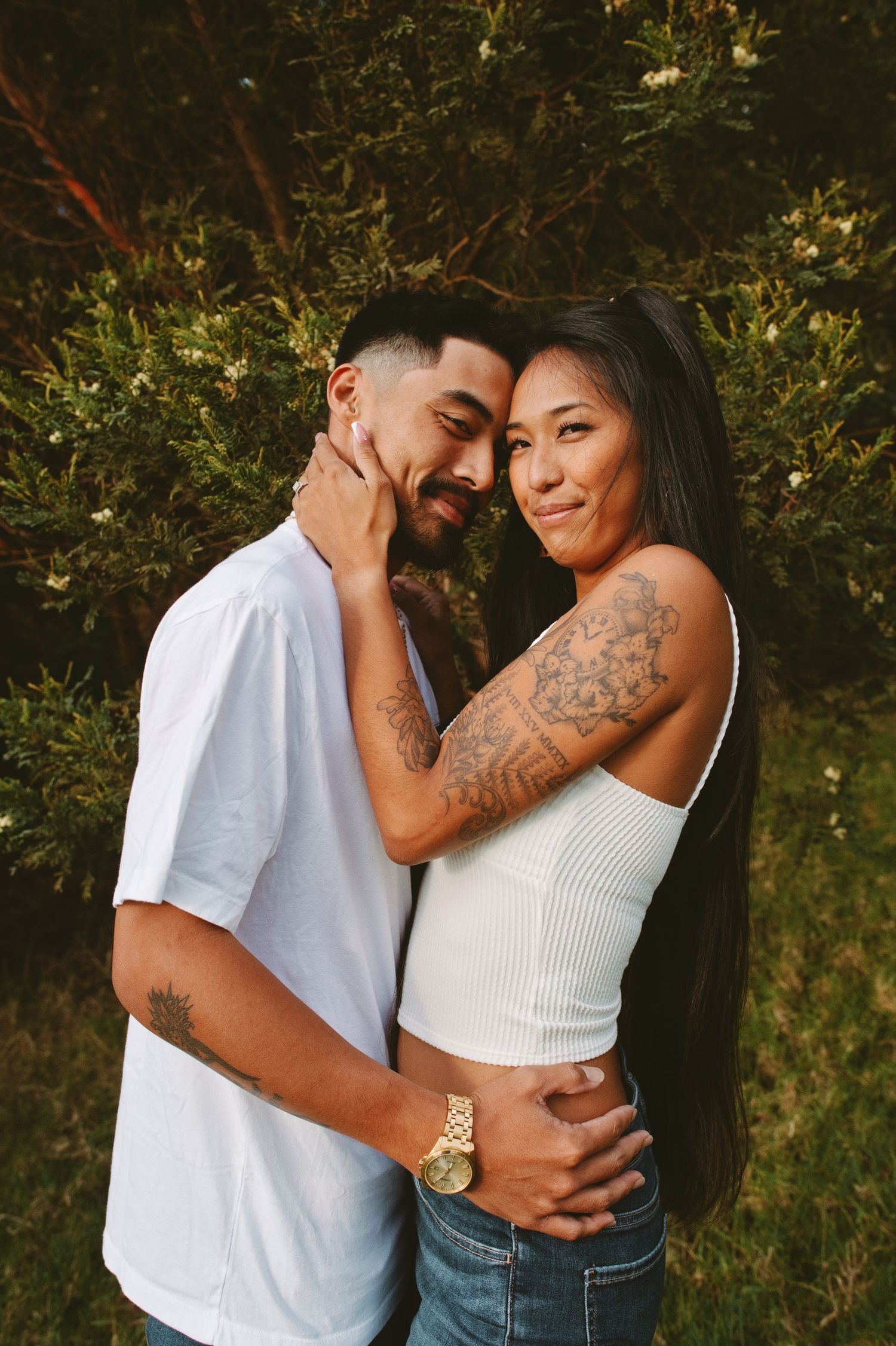 Couple embracing tenderly in front of green bushes, woman with floral tattoo smiling warmly.