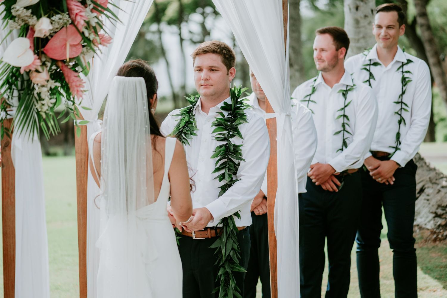 Wedding party members stand under a white draped arch decorated with tropical greenery.