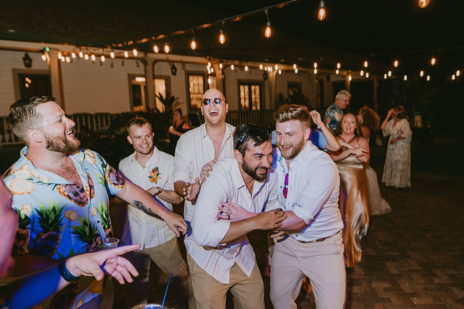 Wedding guests dance and celebrate under string lights at an outdoor reception.