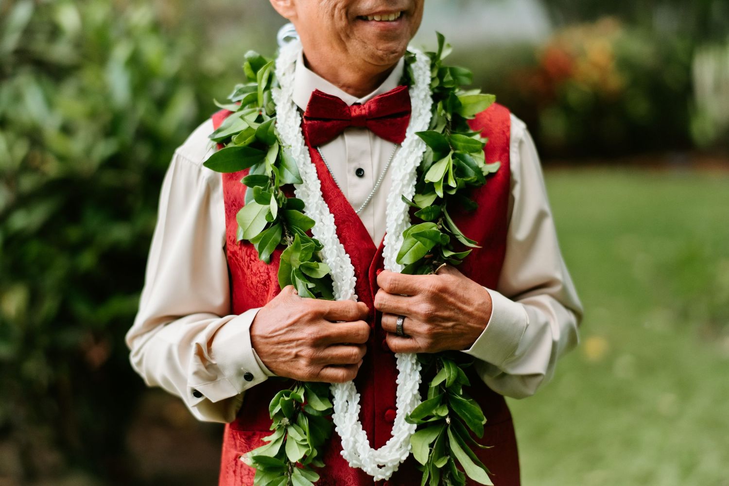 Man in red vest and bow tie wearing traditional Hawaiian lei garlands, smiling outdoors at a garden celebration.