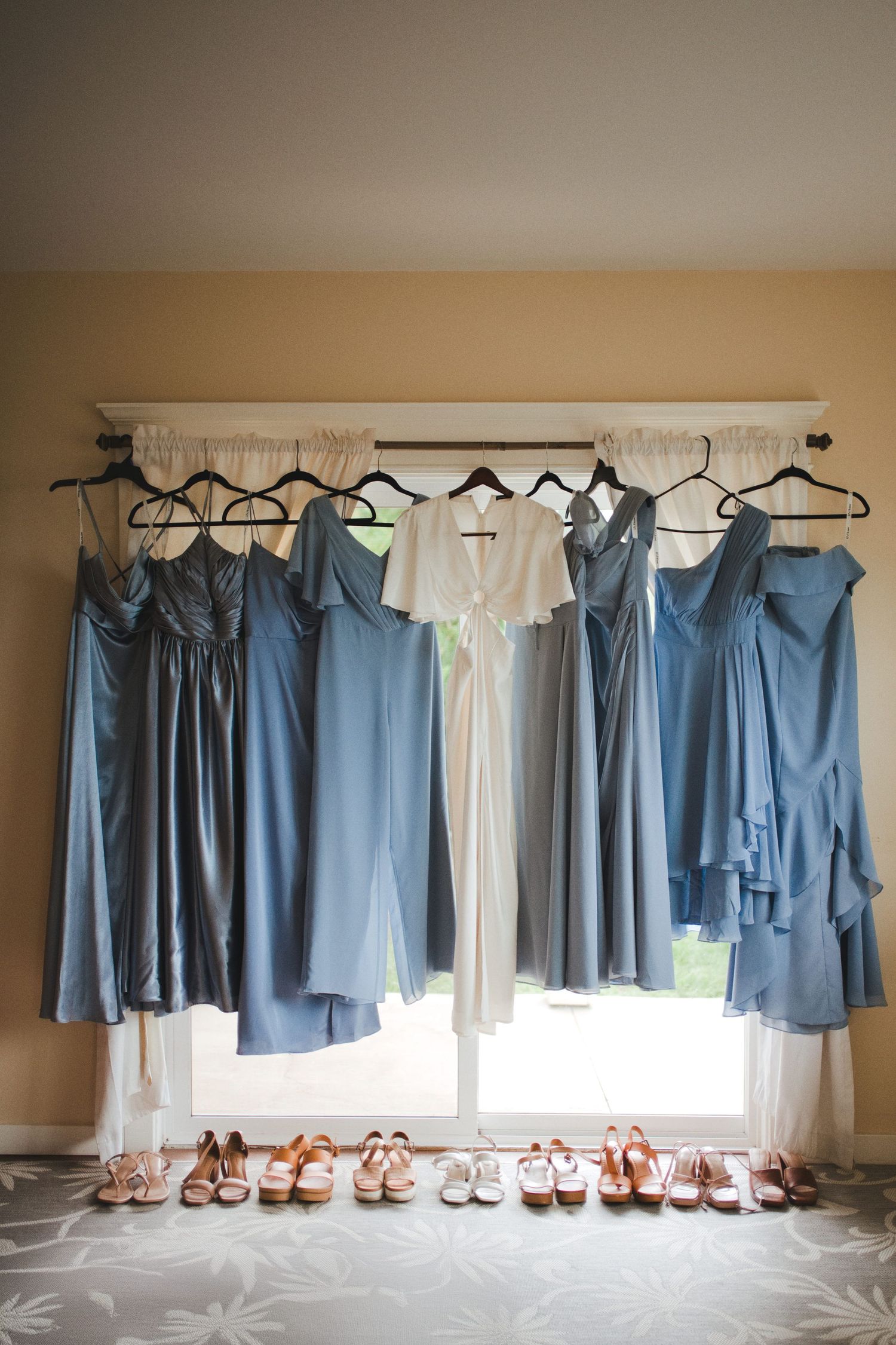 Blue bridesmaid dresses and white bridal gown hanging on a window rod with pink heels arranged below.