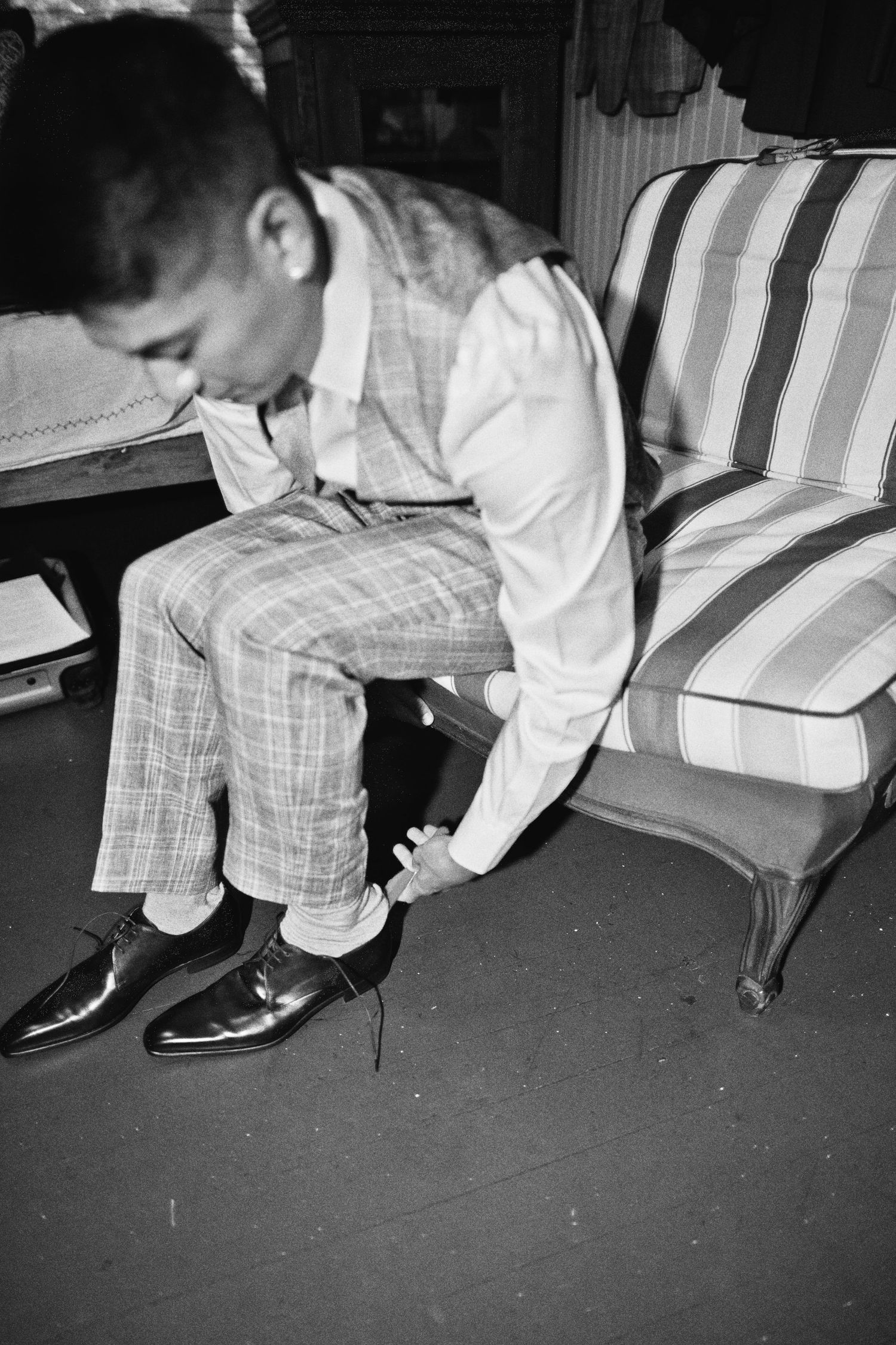 Black and white photo of a man in plaid suit putting on dress shoes while seated on a striped chair.