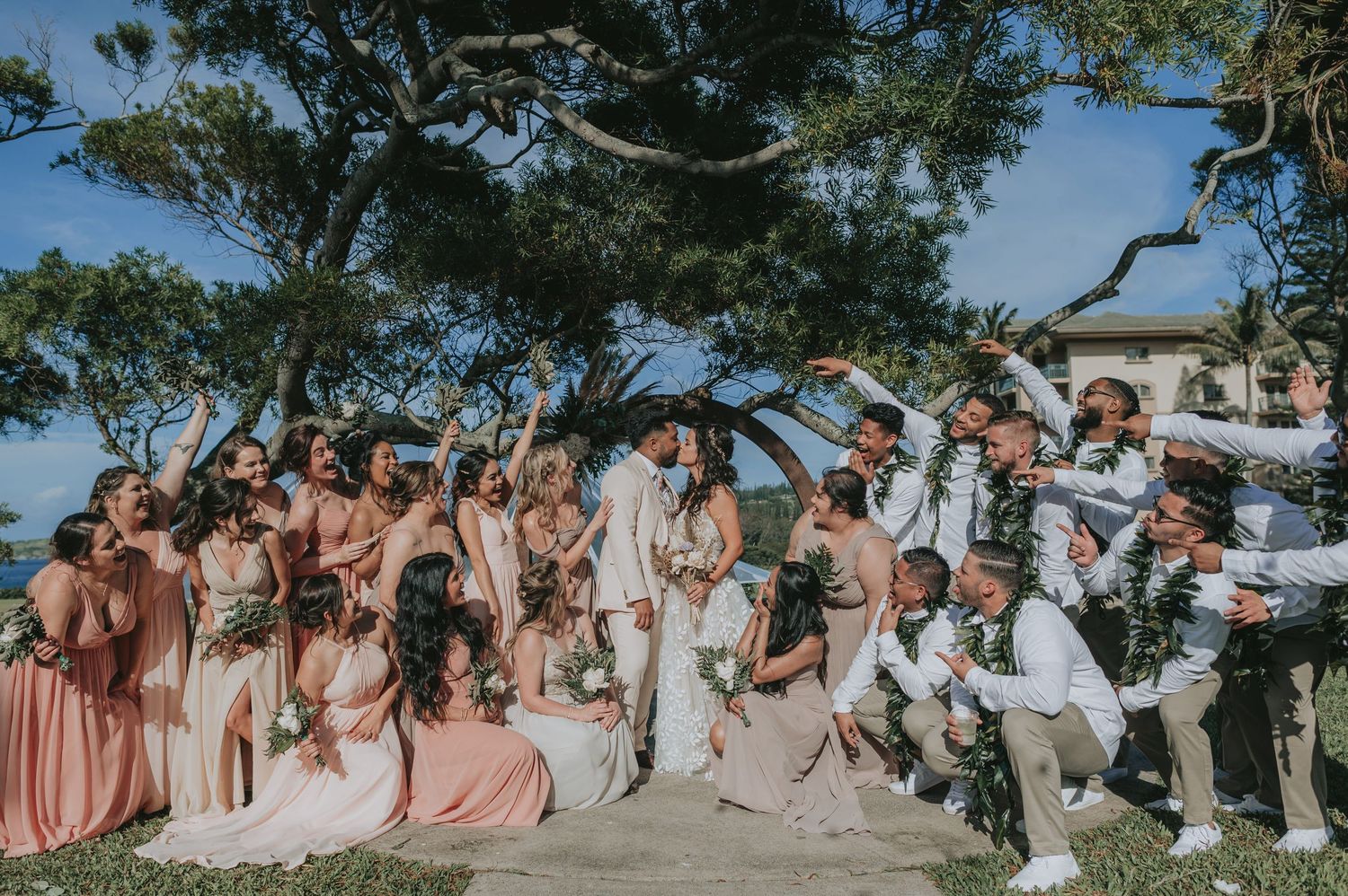 Wedding party celebrating outdoors under trees, bridesmaids in blush pink dresses and groomsmen in white suits cheering.