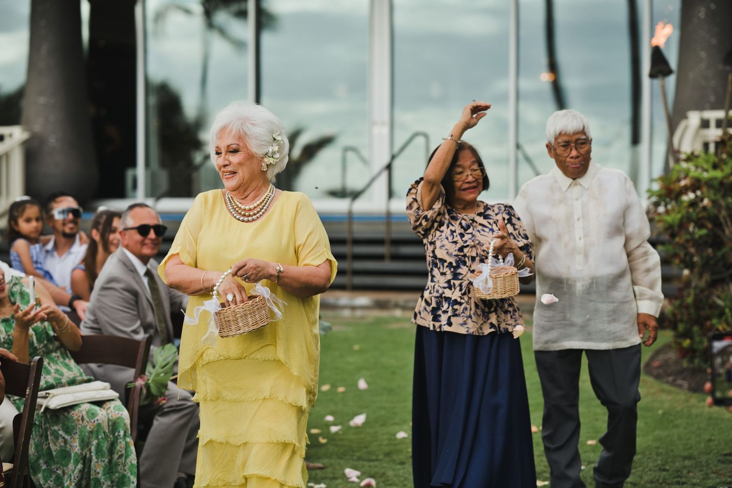 Elegantly dressed older couples walk down the aisle at an outdoor wedding ceremony on a lush green lawn.